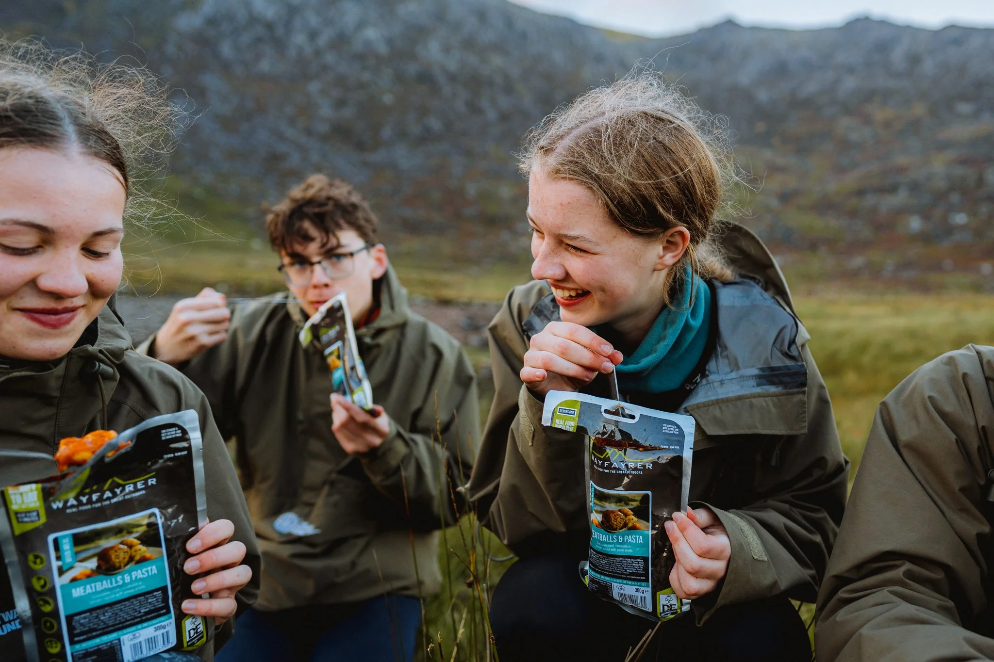 Freelance outdoor Lifestyle Photographer Group of four young people outdoors holding packaged food snacks, smiling and appearing to enjoy a meal during a hike in a scenic mountainous area.