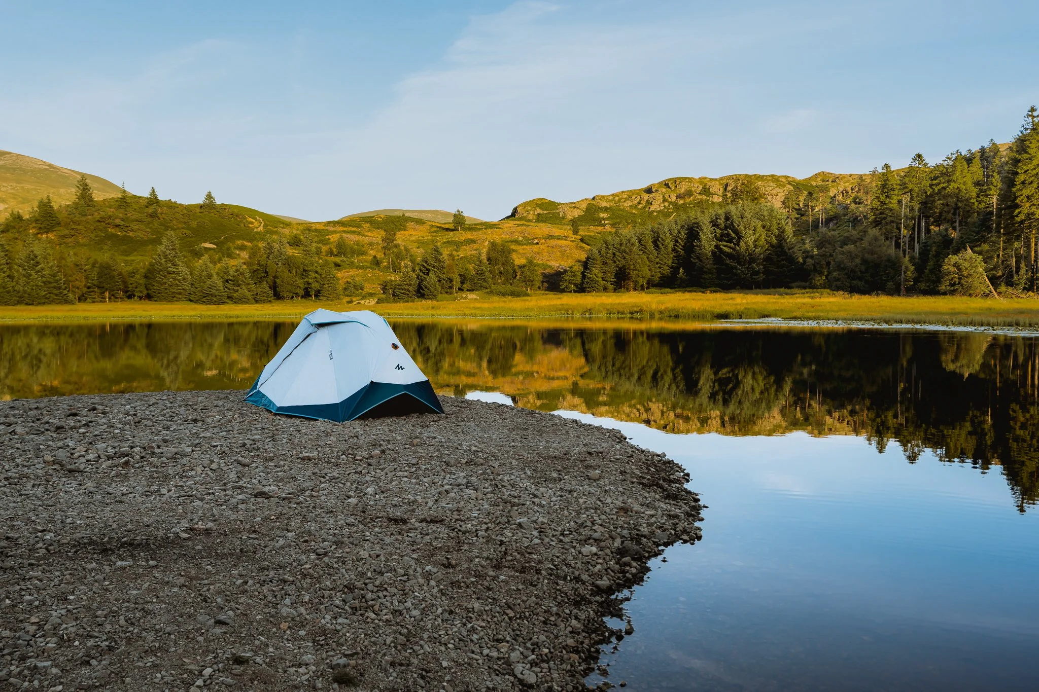 A blue and gray tent set up on a rocky shoreline by a calm lake with mountains and trees in the background, under a clear blue sky.