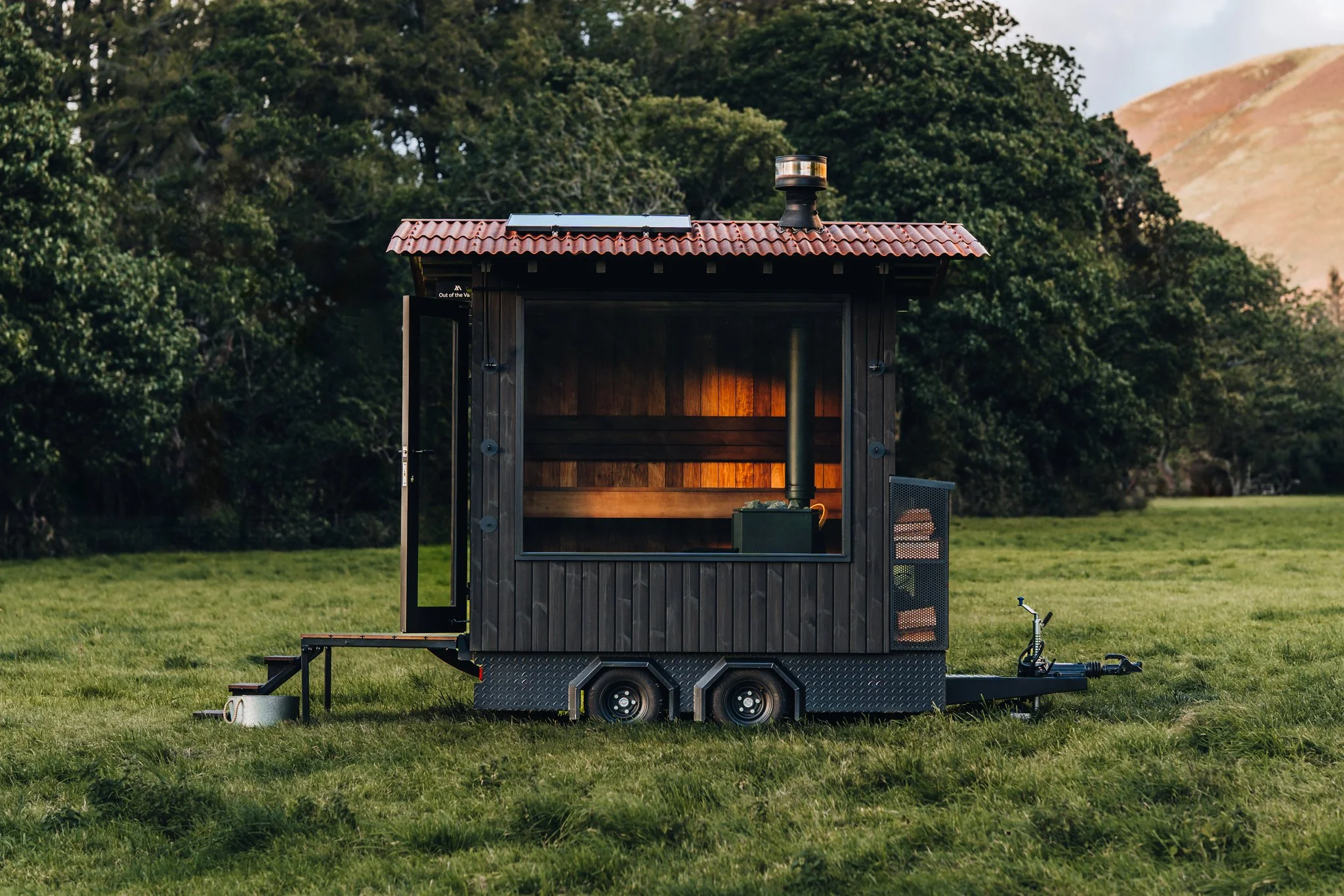 A tiny black mobile cabin on wheels in a grassy field, with open door, inside a small stove and a lit wall heater, backed by trees and distant hills.