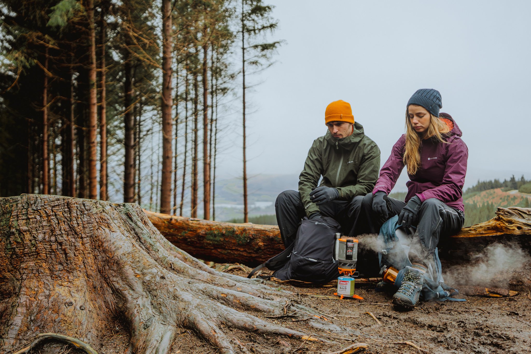 Two hikers, a man and a woman, sitting on a fallen tree trunk in a forest, wearing outdoor gear and warm hats. The woman is tending to a portable stove, which is emitting steam, while the man looks on. There are trees in the background and the weathe