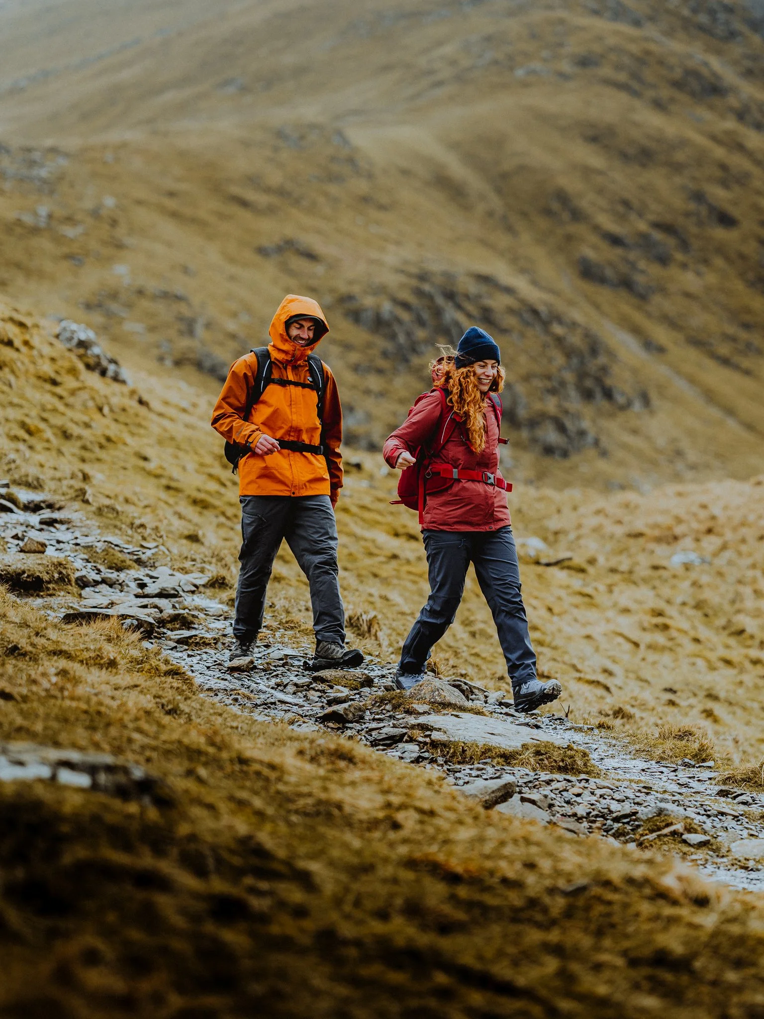 Two hikers walking on a rocky trail in a mountainous landscape, dressed in outdoor gear and smiling.