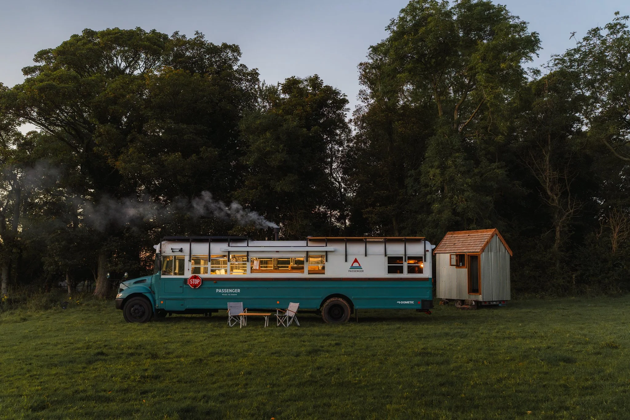 A converted bus parked on a grassy field with a small metal tiny house on wheels nearby, trees in the background, and outdoor chairs in front of the bus.