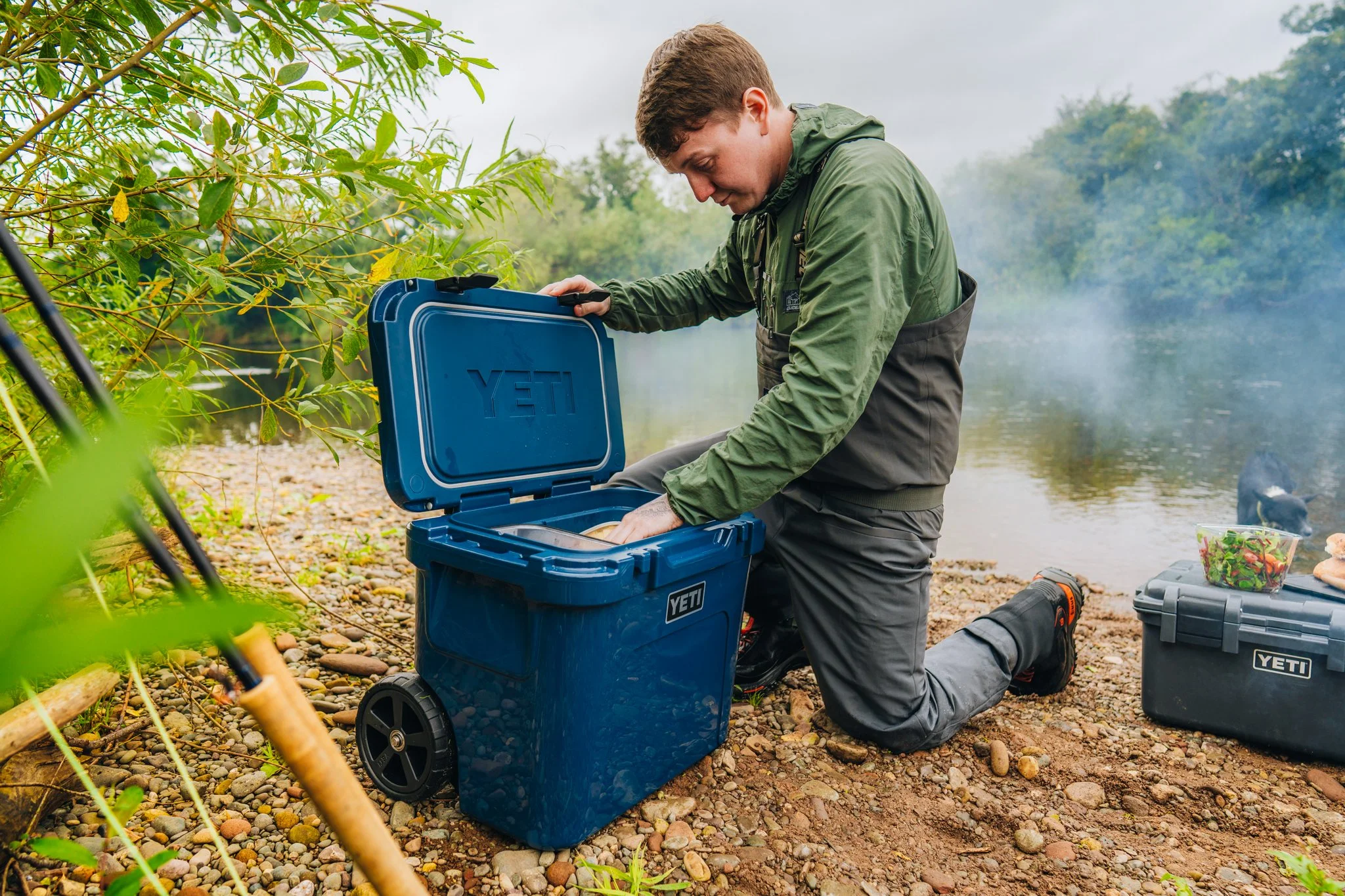 A man kneels on the rocky ground near a river by the woods, opening a blue YETI cooler with items inside, with a second YETI cooler and a salad bowl on the ground nearby.