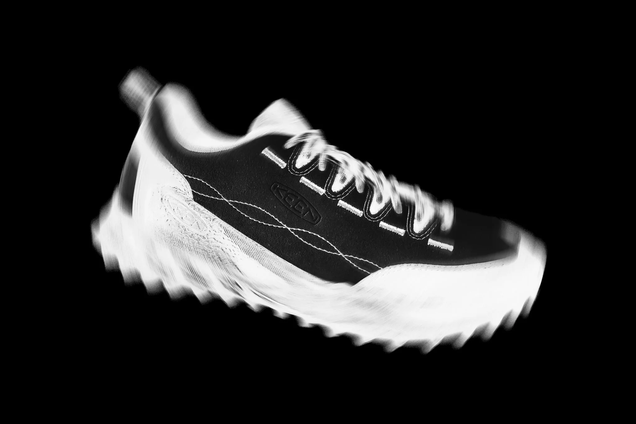 Freelance outdoor Lifestyle Photographer Black and white image of a running shoe with a thick, textured sole, displayed against a dark background. KEEN 