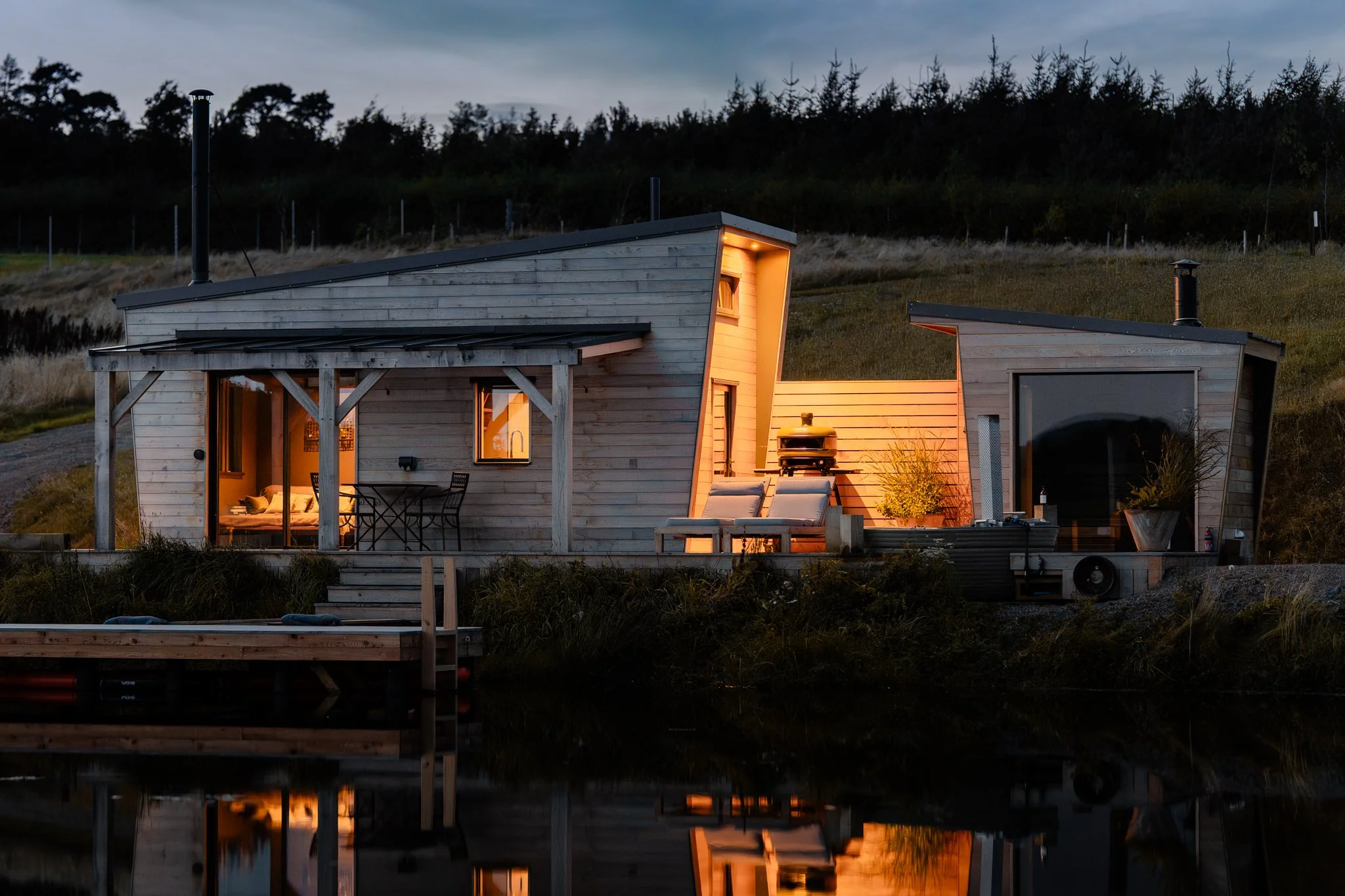Modern wooden house illuminated at dusk, with outdoor lounge chairs and a small deck near water, surrounded by grassy landscape and trees in the background.