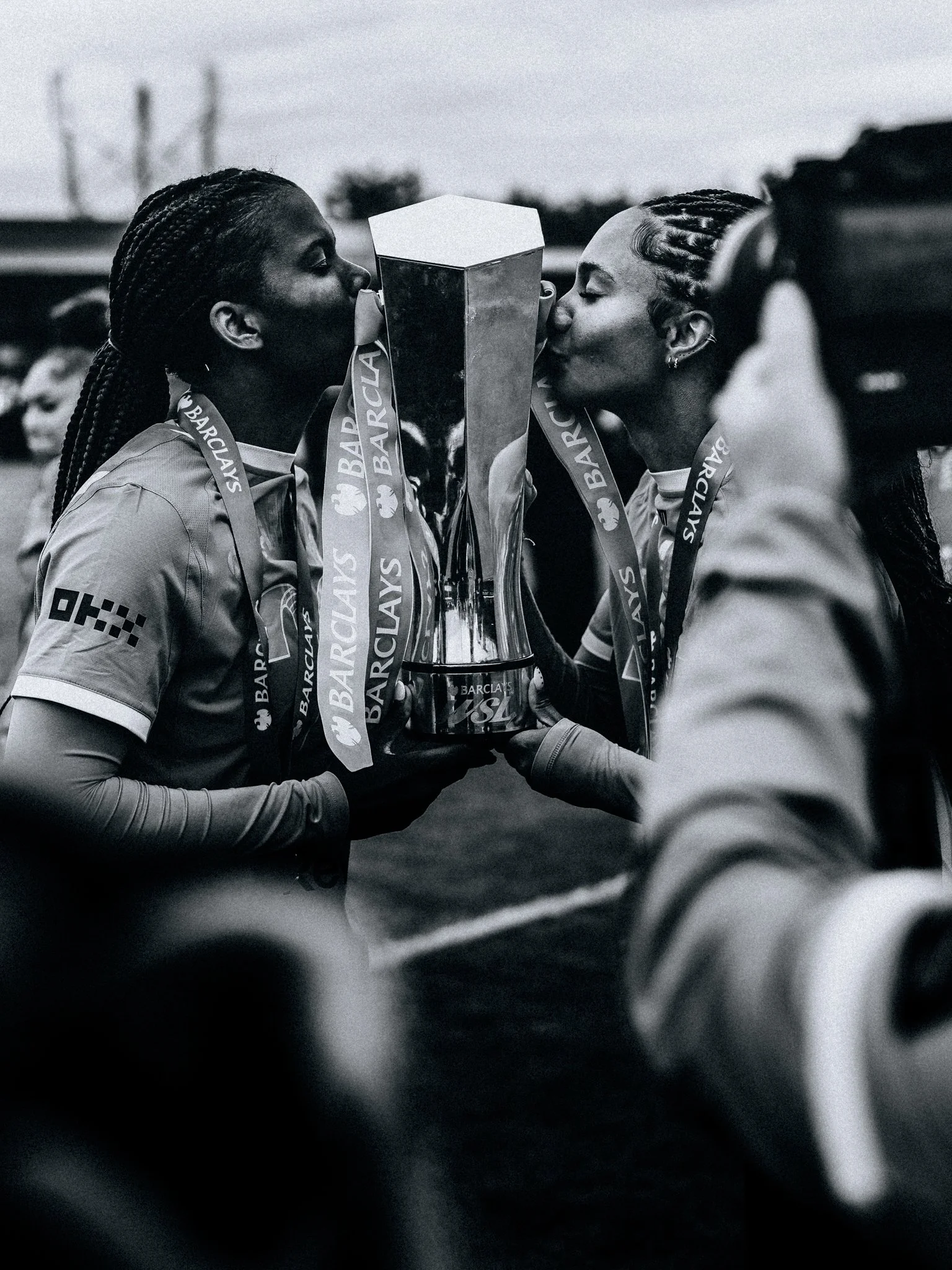 Two women in athletic gear kissing and holding a large trophy with medals around their necks, celebrating a victory.