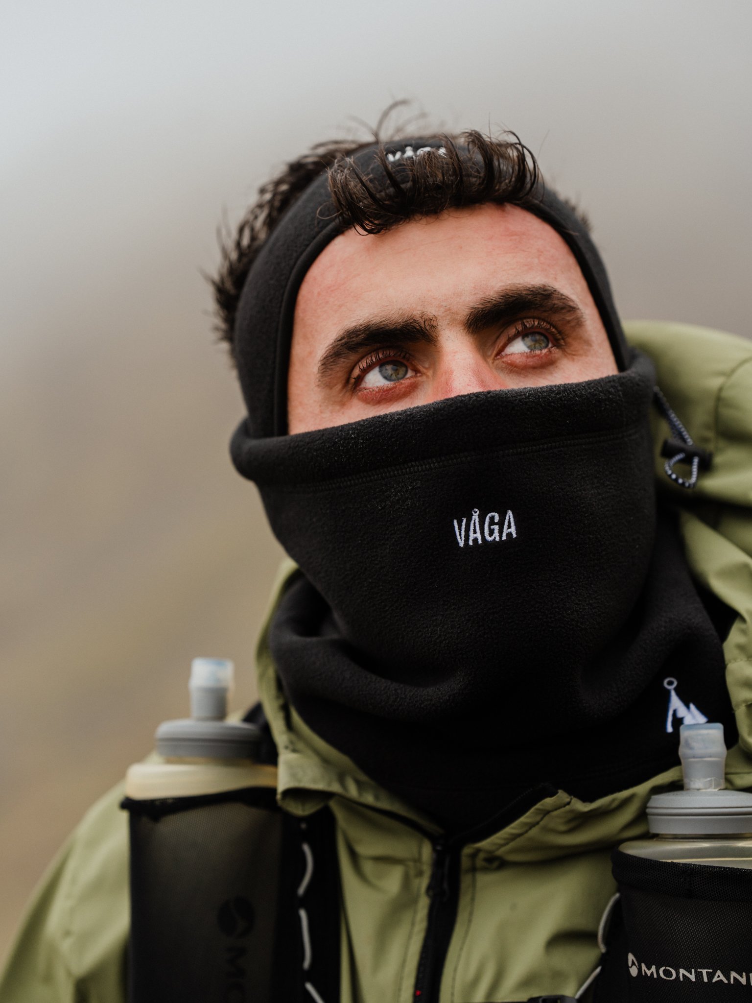 Freelance outdoor Lifestyle Photographer Close-up of a man wearing a black face mask with the word 'VÁGA' on it, a black headband, and outdoor gear, looking up against a cloudy sky.