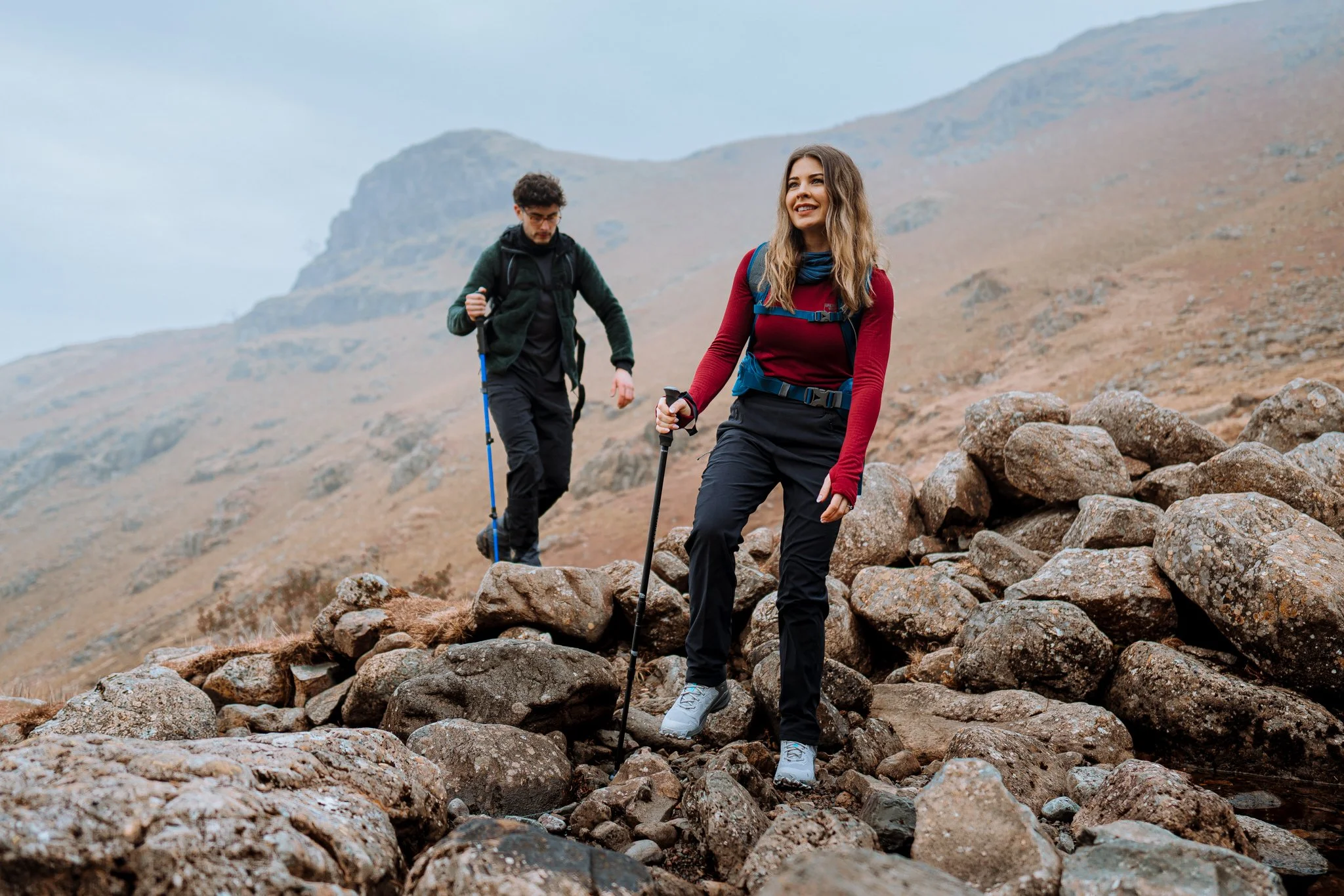 Freelance Lifestyle Photographer Two hikers climbing over rocks on a mountain trail with a hilly landscape in the background, one woman in a red sweater and black pants leading, man behind in black clothing.