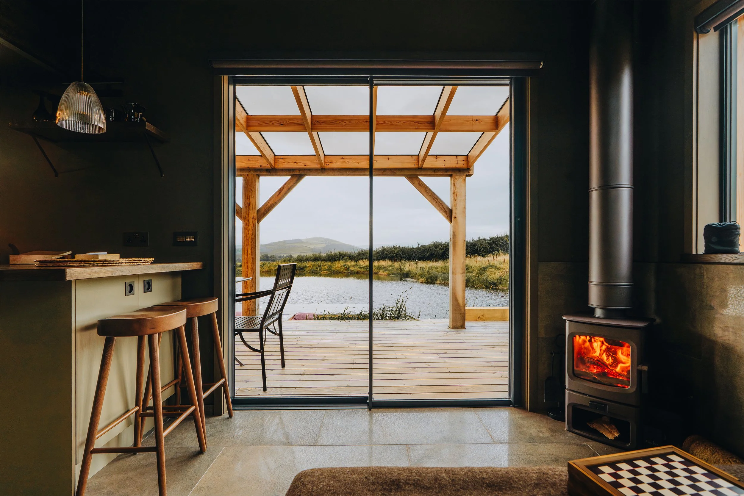 Interior of a cozy cabin with a wood stove and a view of a lake and mountains outside through sliding glass doors.
