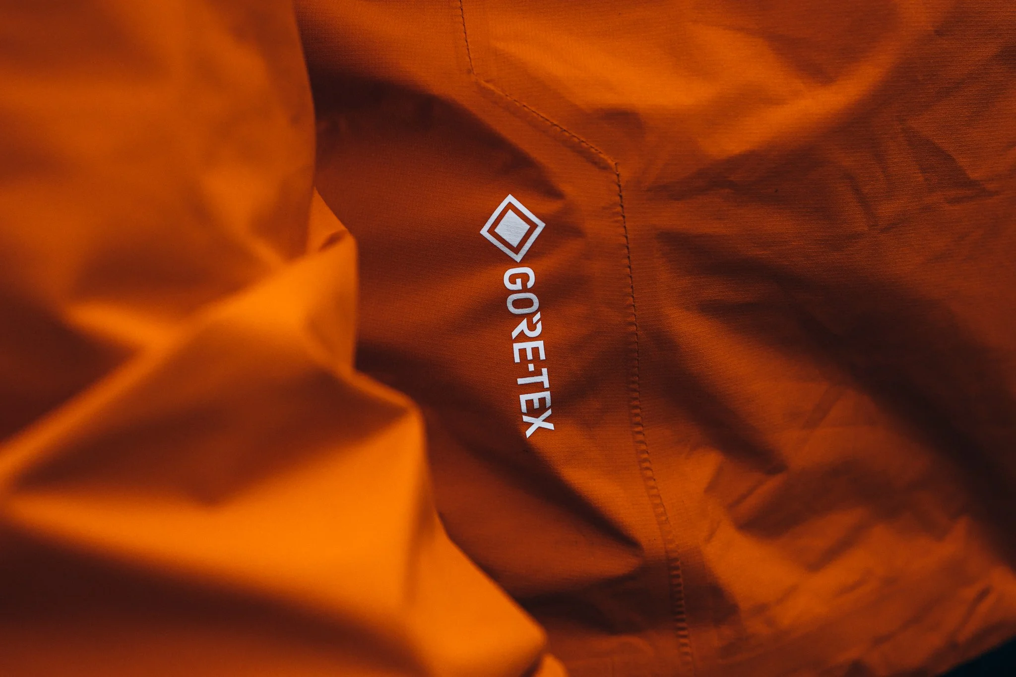 Close-up of orange outdoor jacket interior with white text and logo reading 'GORE-TEX' printed on the fabric.