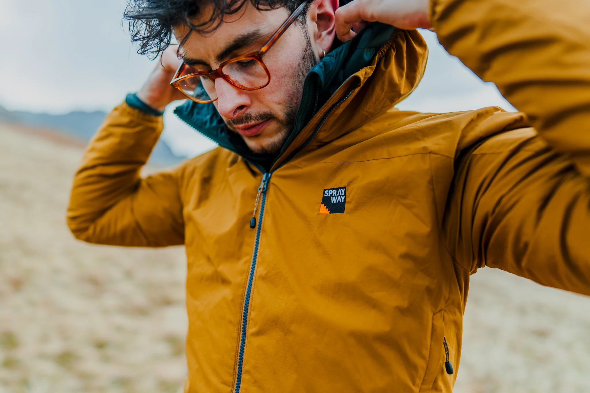 Freelance outdoor Lifestyle Photographer A man wearing glasses and a yellow jacket with a blue zipper, standing outdoors with mountains in the background, adjusting his hood.