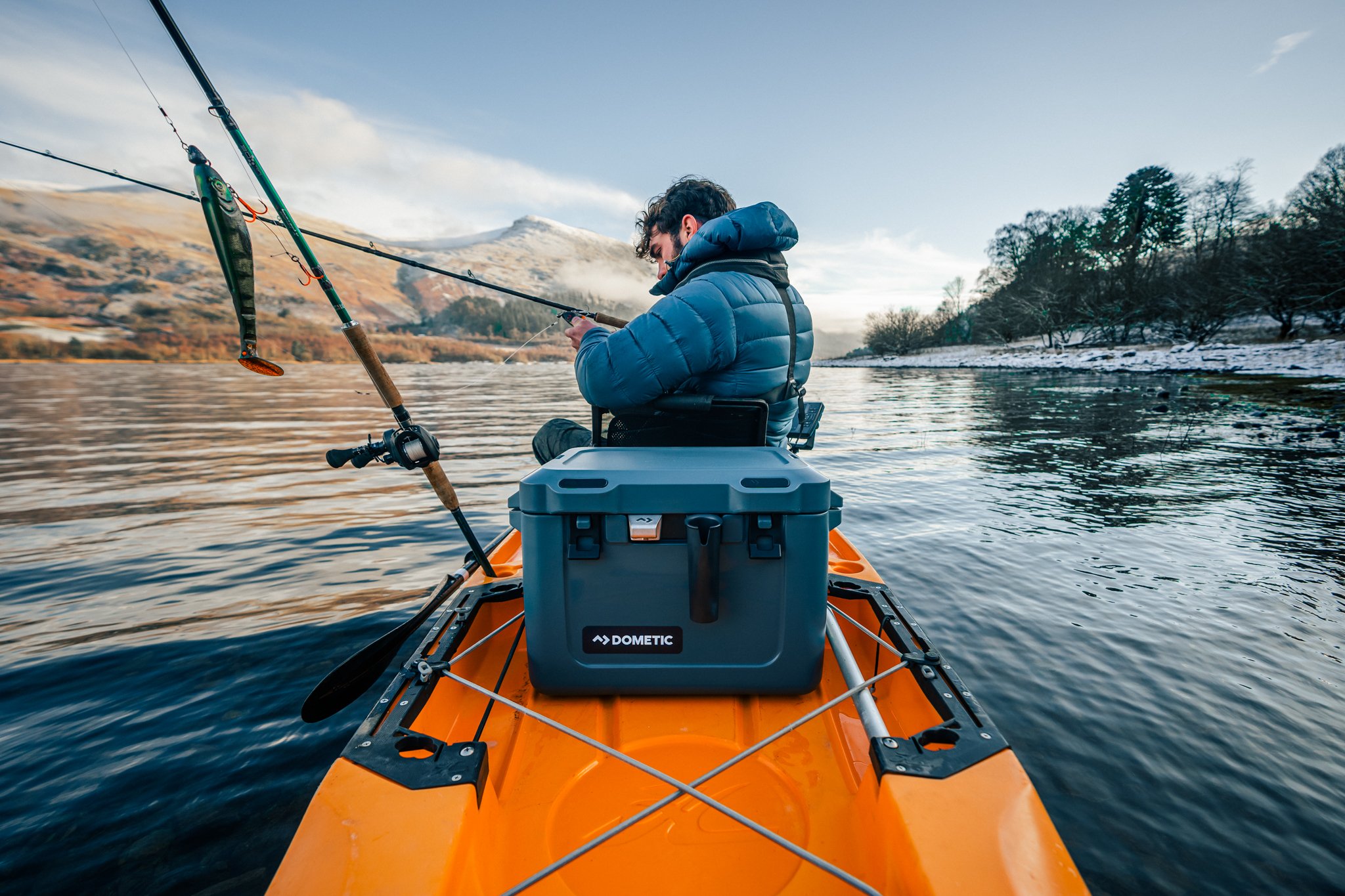 Person in a blue puffer jacket sitting in an orange kayak, fishing in a lake with mountains and trees in the background.