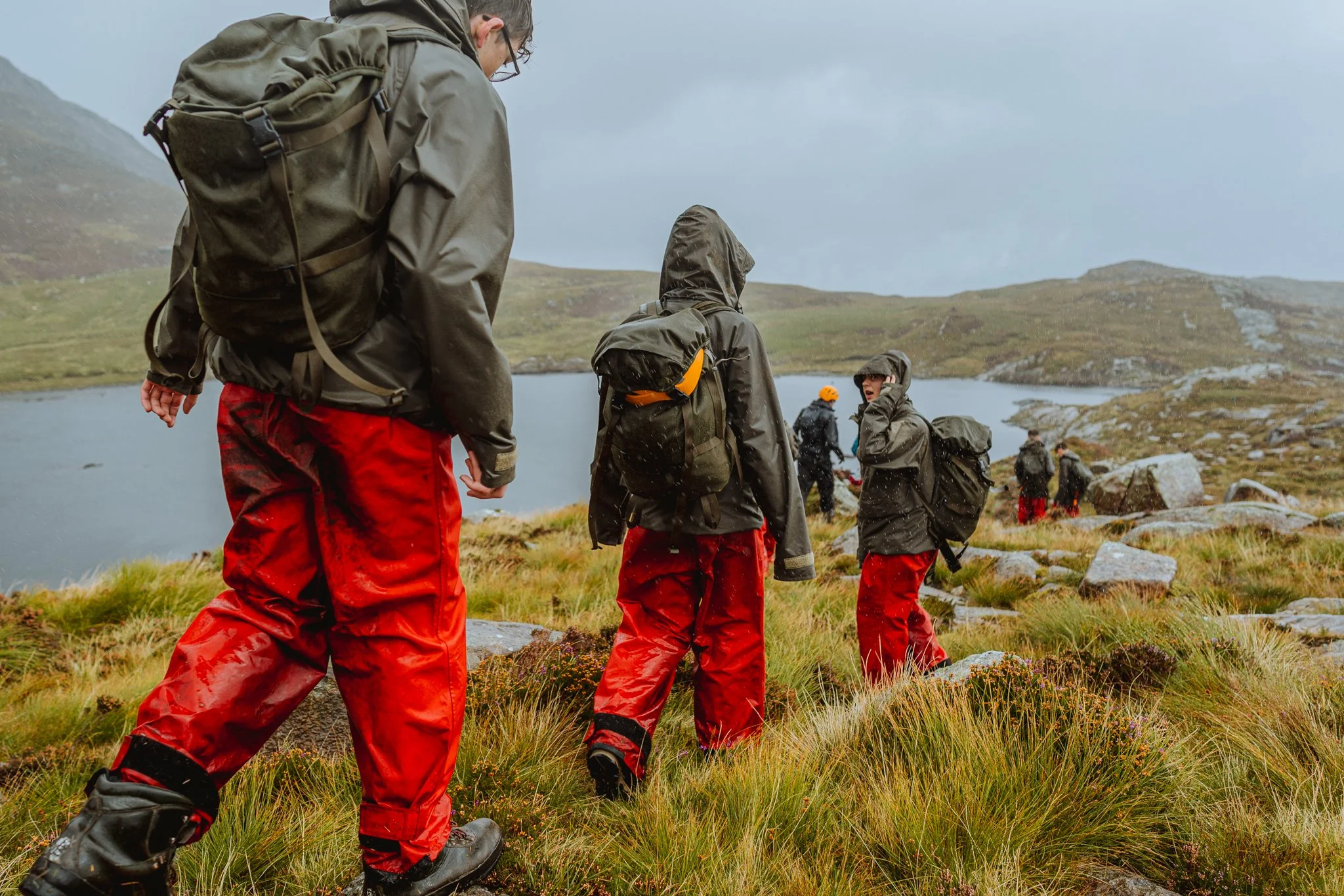 A group of people wearing outdoor gear, red waterproof pants, and backpacks hiking near a lake in a hilly, grassy landscape with rocks and cloudy sky.