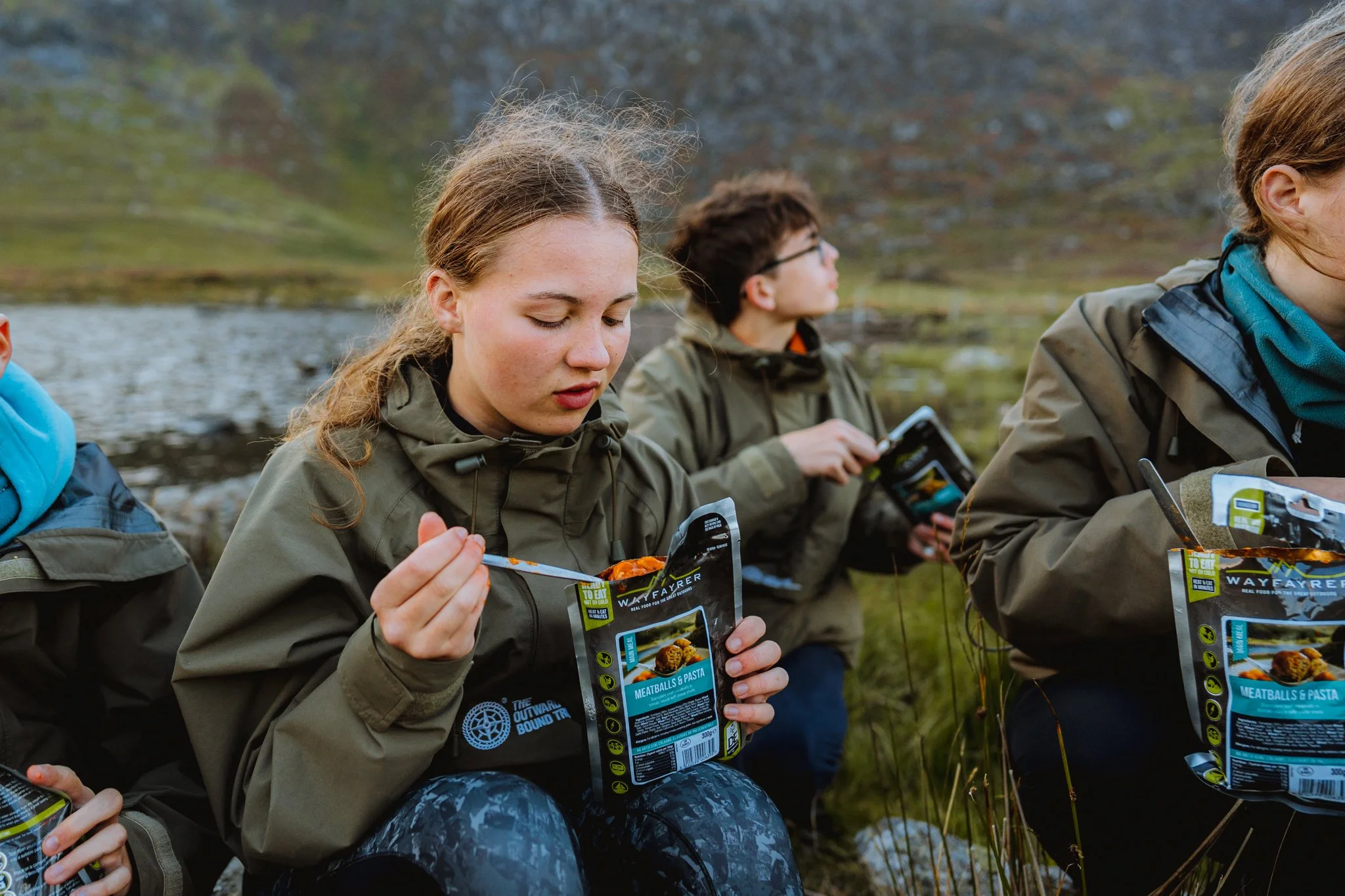 Freelance outdoor Lifestyle Photographer Group of young people in outdoor gear sitting on grass near a lake, eating packaged meal with spoons.