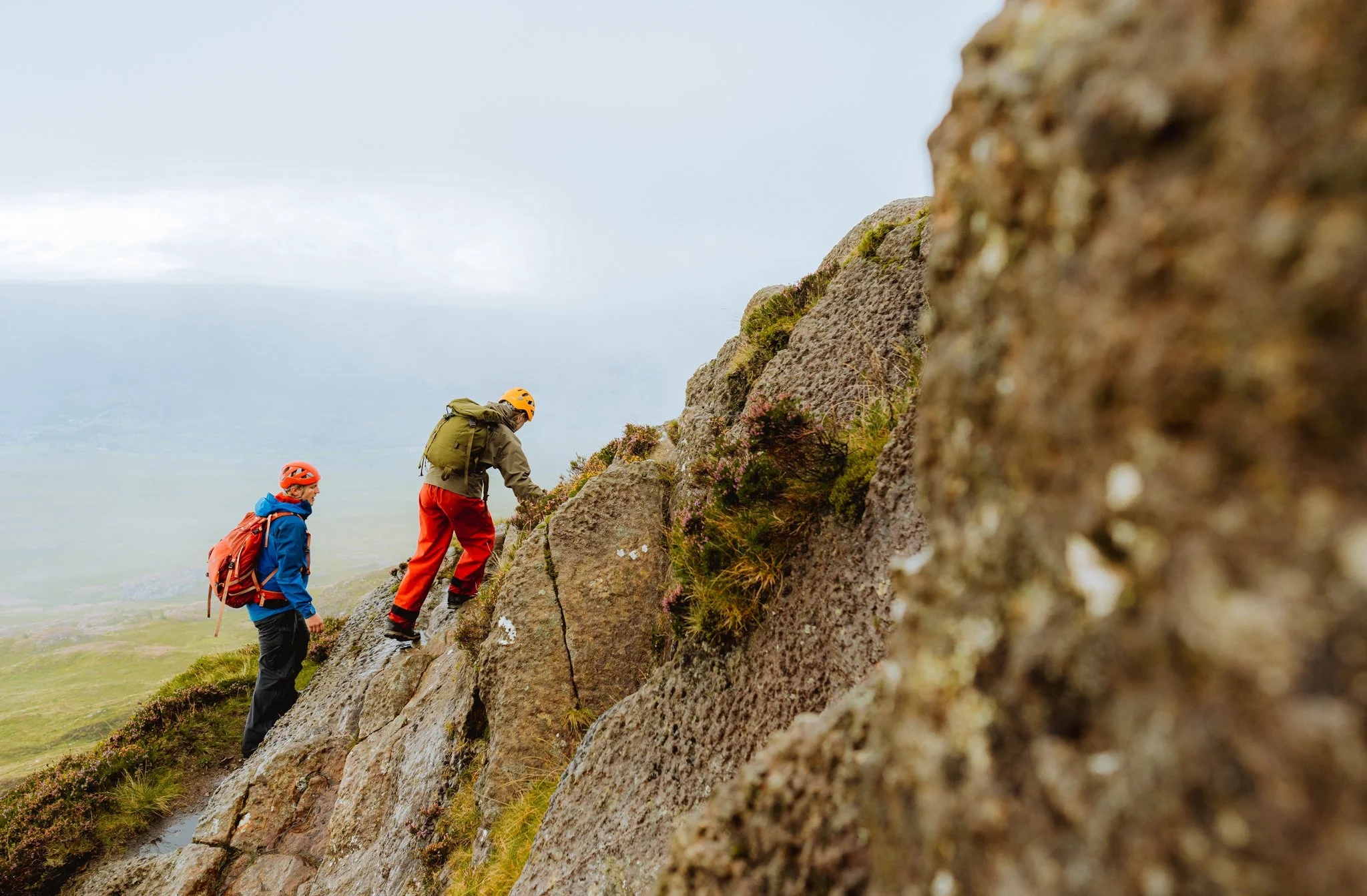 Two climbers with backpacks and helmets ascending a steep rocky mountain trail with green grass and plants, under a cloudy sky.