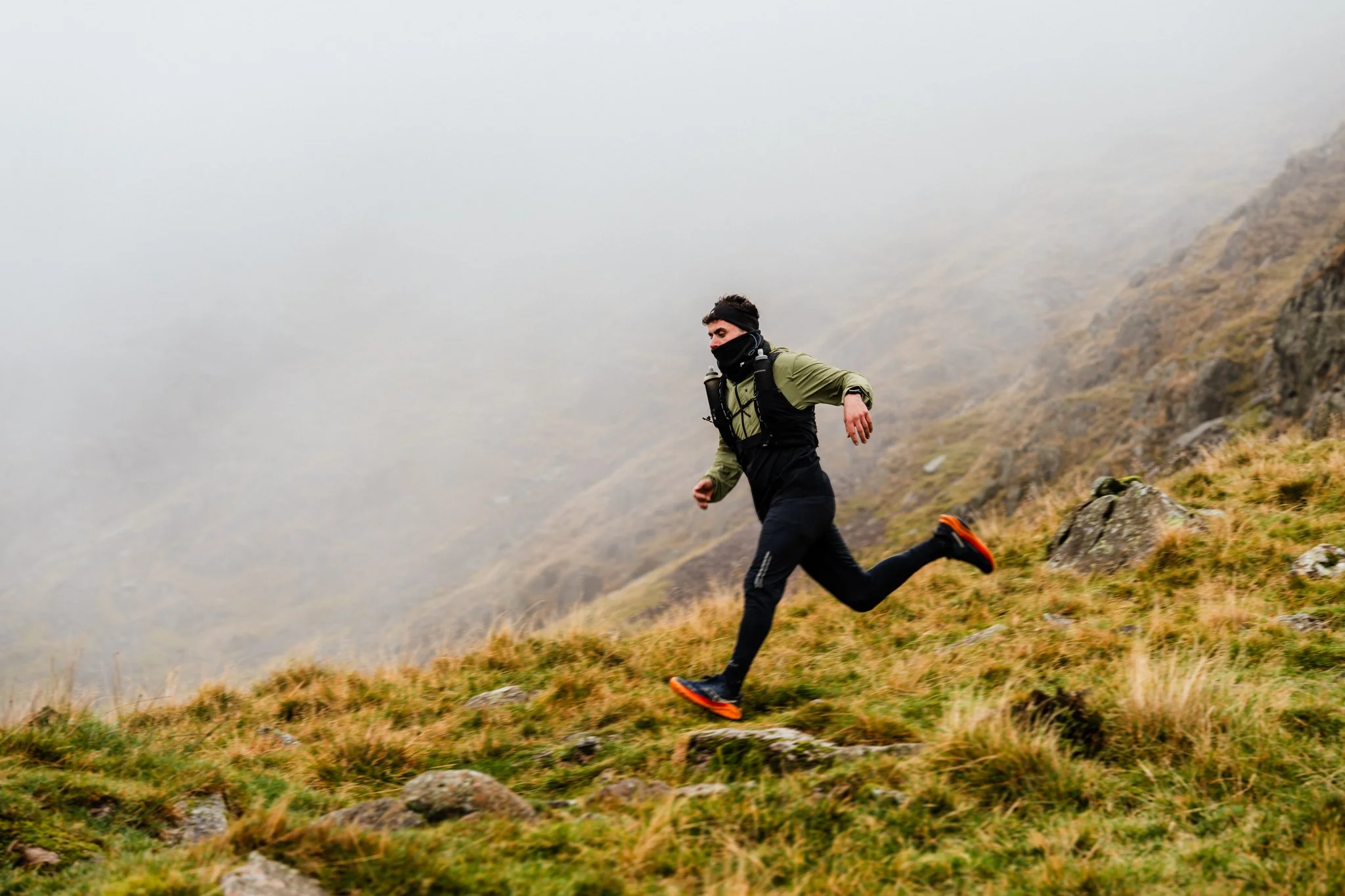A man running on a foggy mountain trail with rocks and grass, wearing outdoor gear and a backpack.
