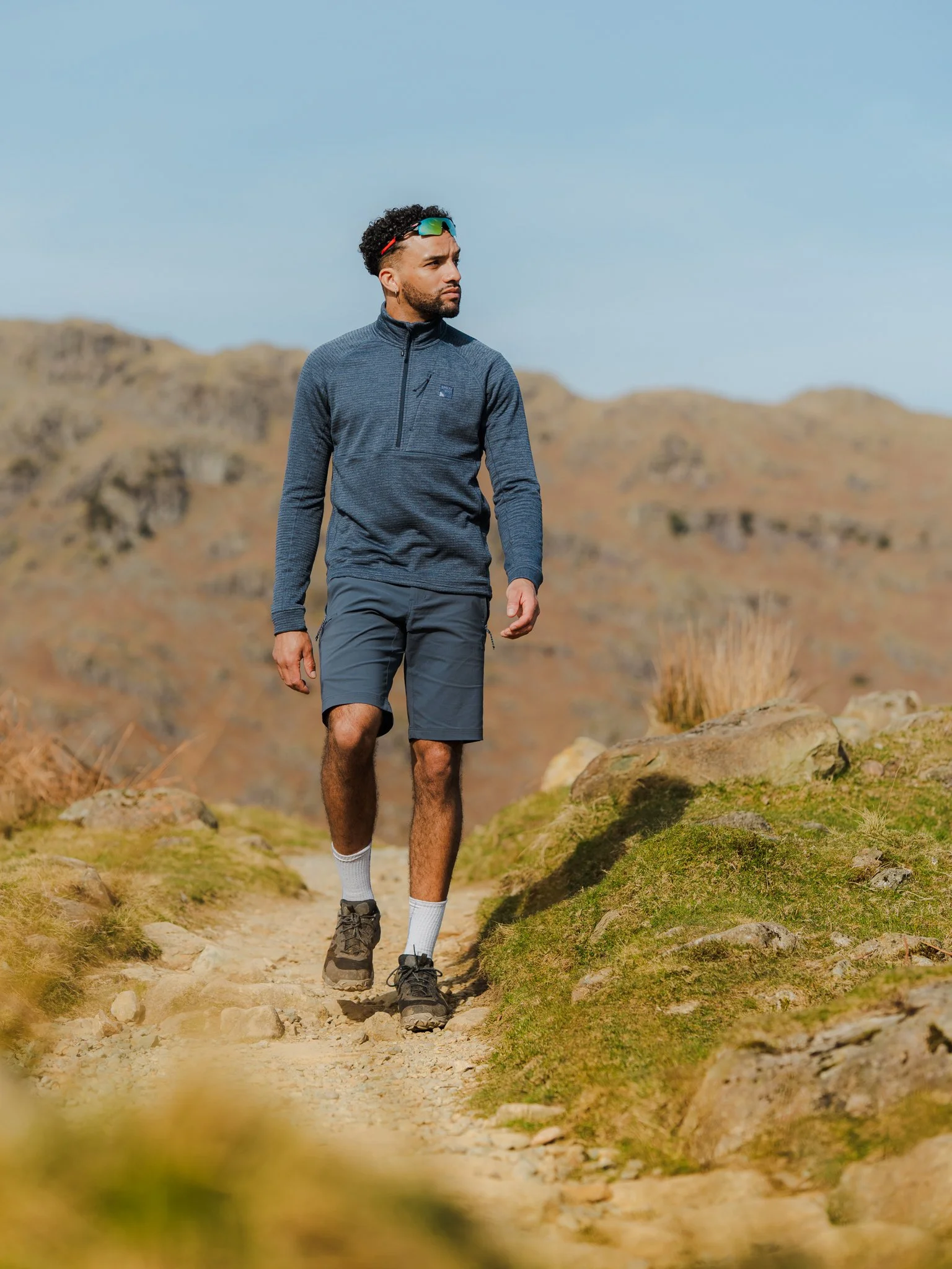 Freelance Lifestyle Photographer A man walking on a rocky trail in a mountainous area with dry grass and hills in the background, wearing a dark blue athletic jacket, shorts, and hiking shoes.