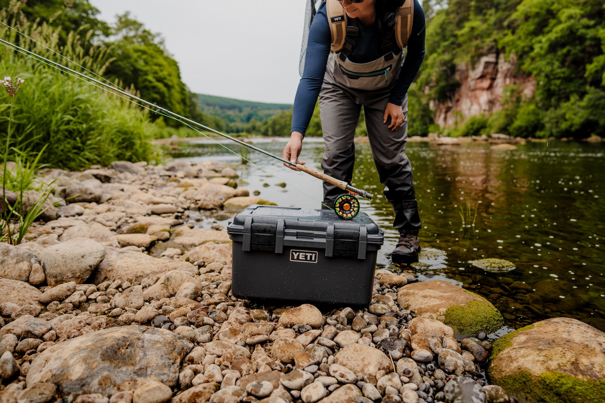 Freelance outdoor Lifestyle Photographer Person fishing with a rod near a river, using a YETI cooler on rocks at the riverbank, surrounded by green trees and rocks in a scenic outdoor setting.