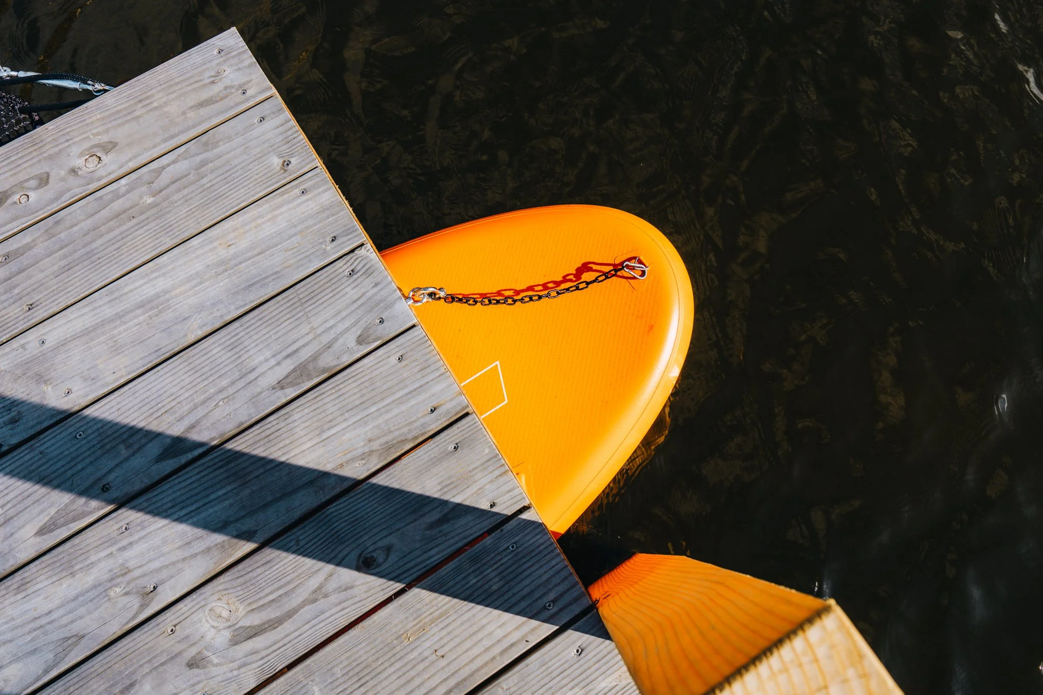 Freelance outdoor Lifestyle Photographer View from above of a wooden dock over water, with an orange paddleboard tied to the dock and two orange paddles resting against the dock edge, cast shadow on the dock.