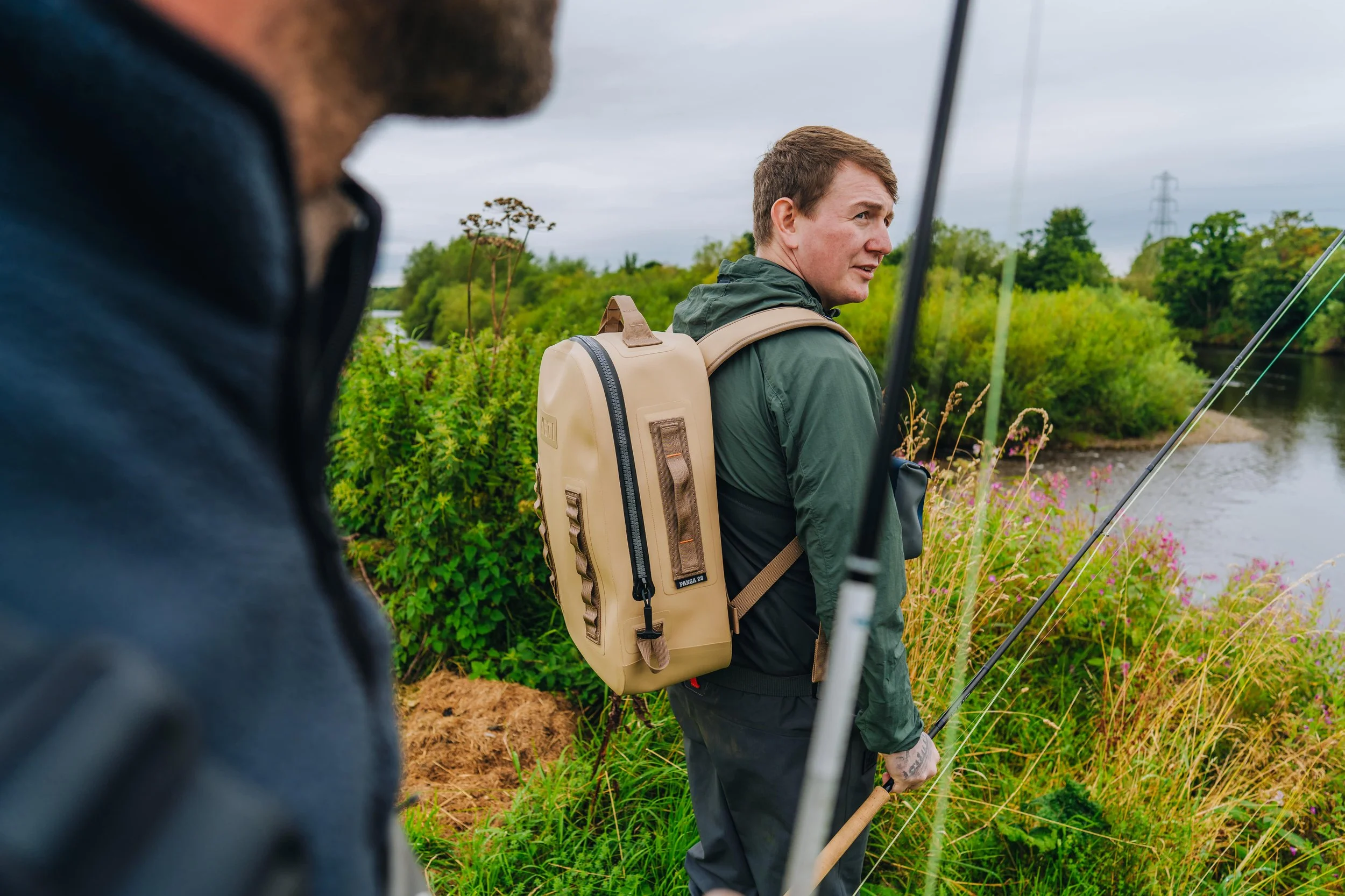 Two men fishing by a river surrounded by greenery, one in the foreground partially out of focus and another in the background holding a fishing rod, wearing a green jacket and backpack.