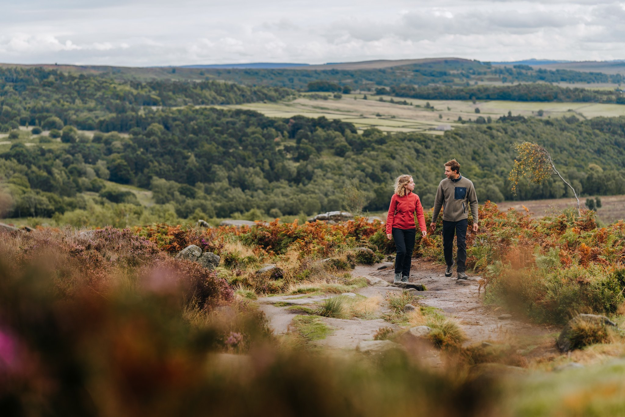 A man and a woman hiking on a trail in a scenic countryside with rolling green hills and trees in the background.