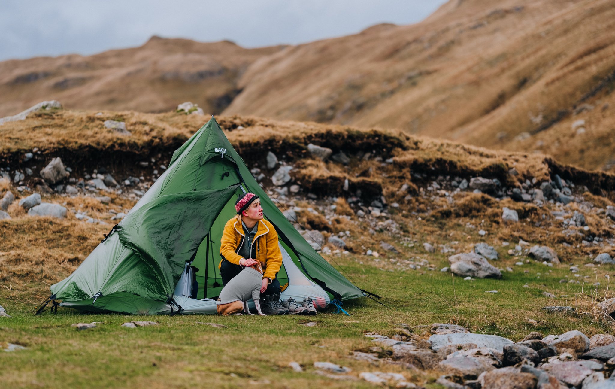 Freelance outdoor Lifestyle Photographer A woman sitting at the entrance of a green camping tent on a grassy hillside with rocky terrain and brown hills in the background, wearing a yellow jacket and a pink hat.
