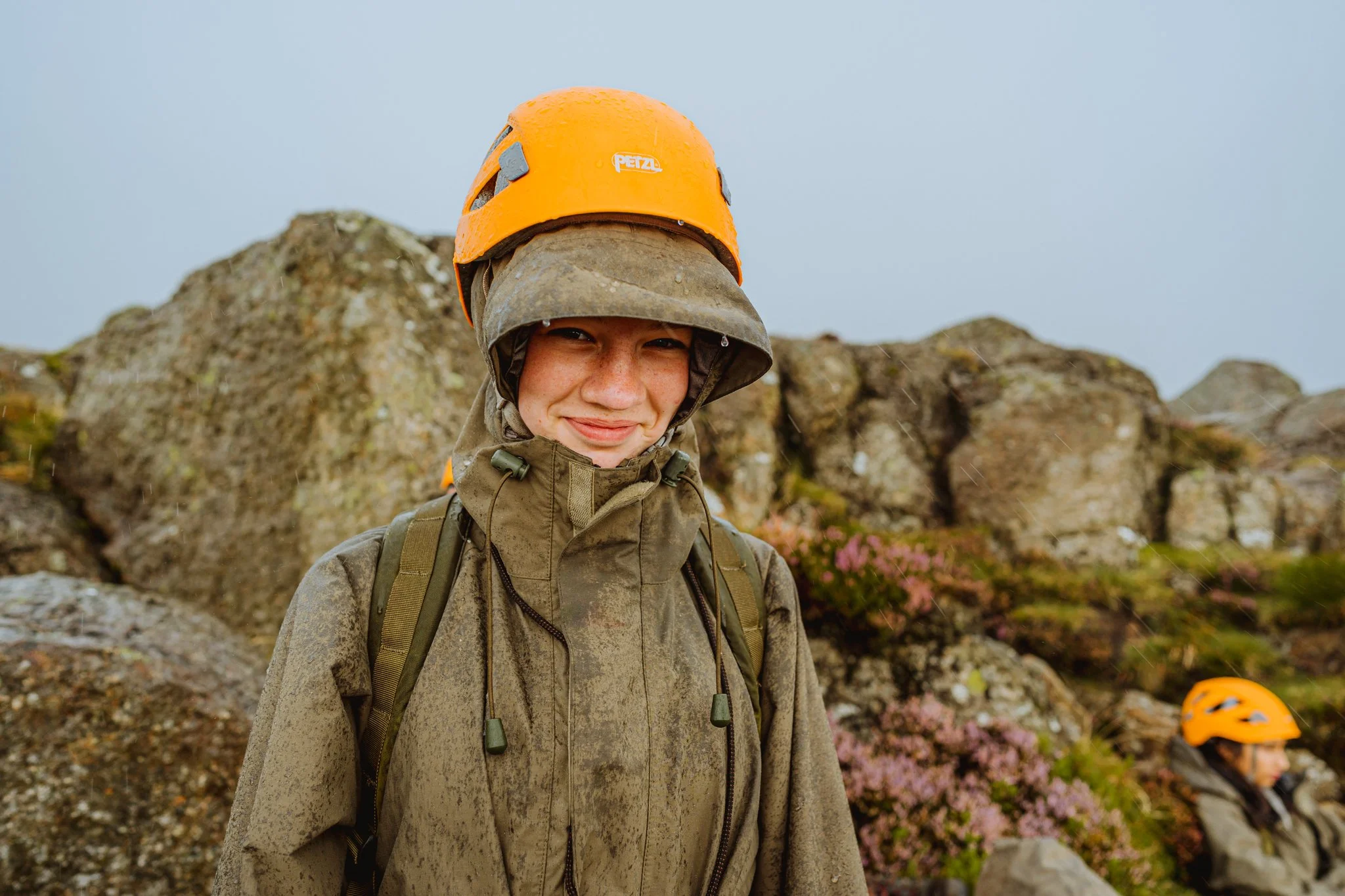 A woman wearing a helmet and rain jacket smiling in front of rocky terrain with pink flowers, another person in helmet and rain gear visible in the background.