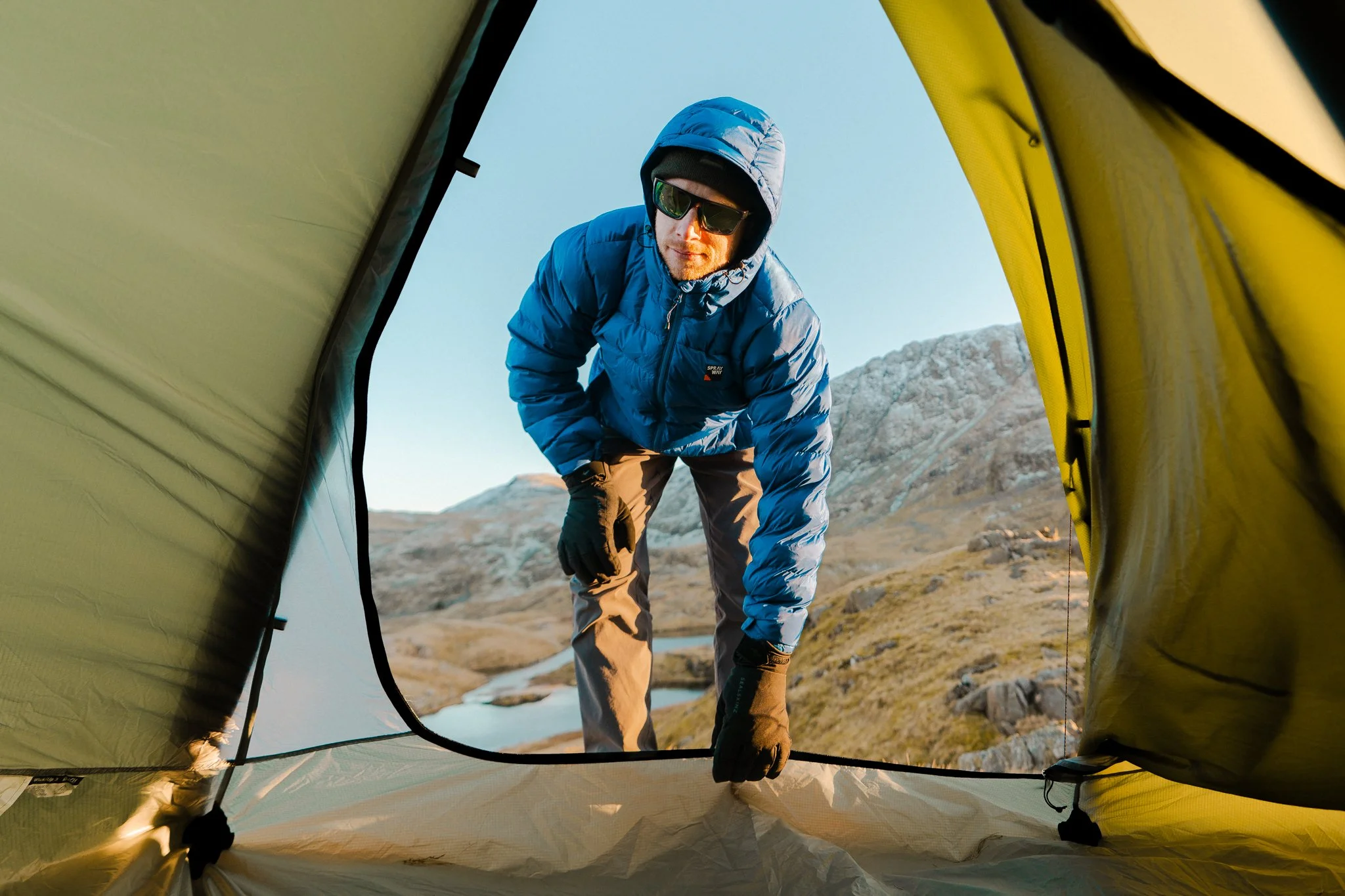 Freelance outdoor Lifestyle Photographer A man dressed in a blue jacket, gray pants, and black gloves is looking down into the camera inside a yellow tent, with a mountainous landscape and river in the background.