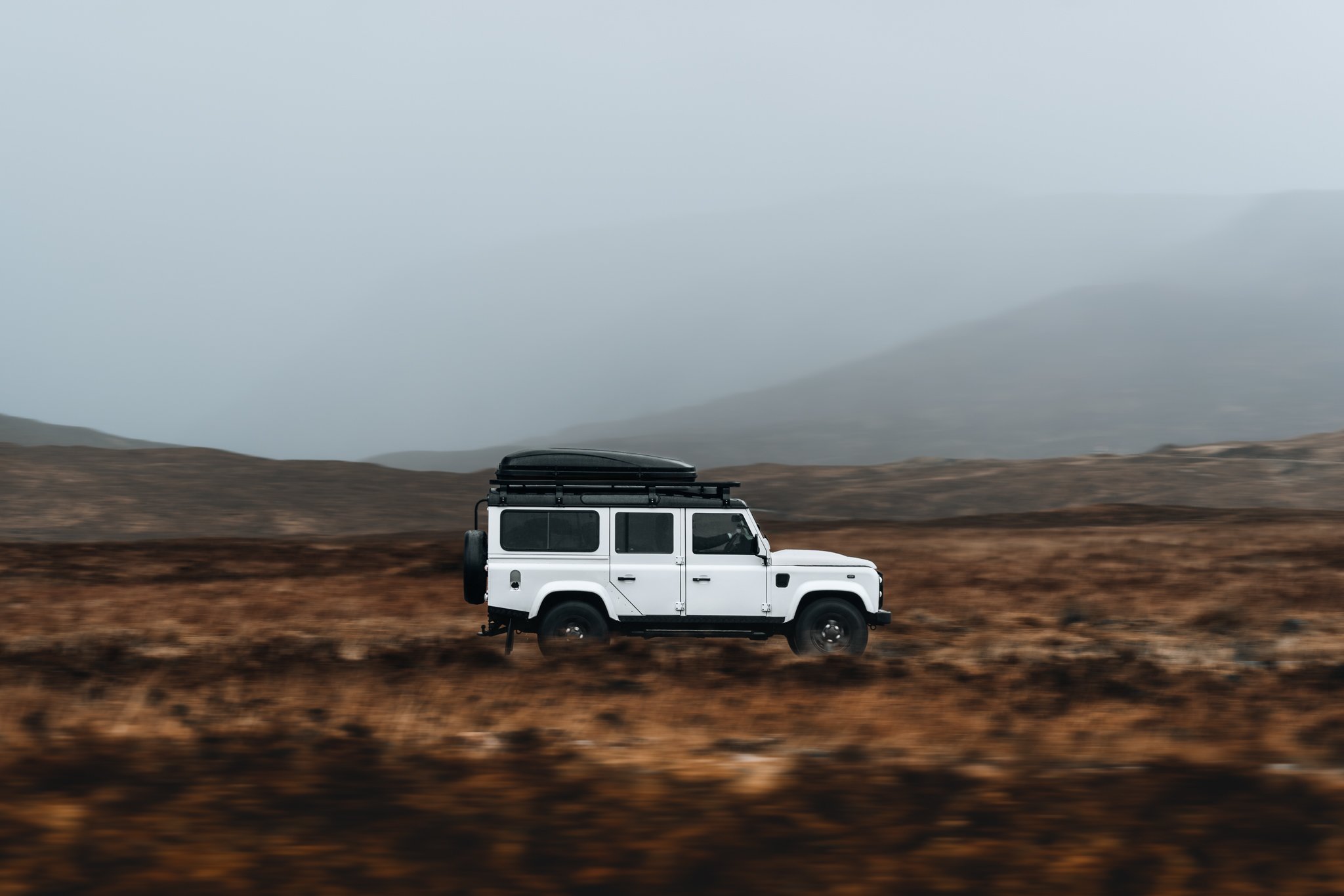A white off-road vehicle driving through a brown, open landscape with mountains and cloudy sky in the background.