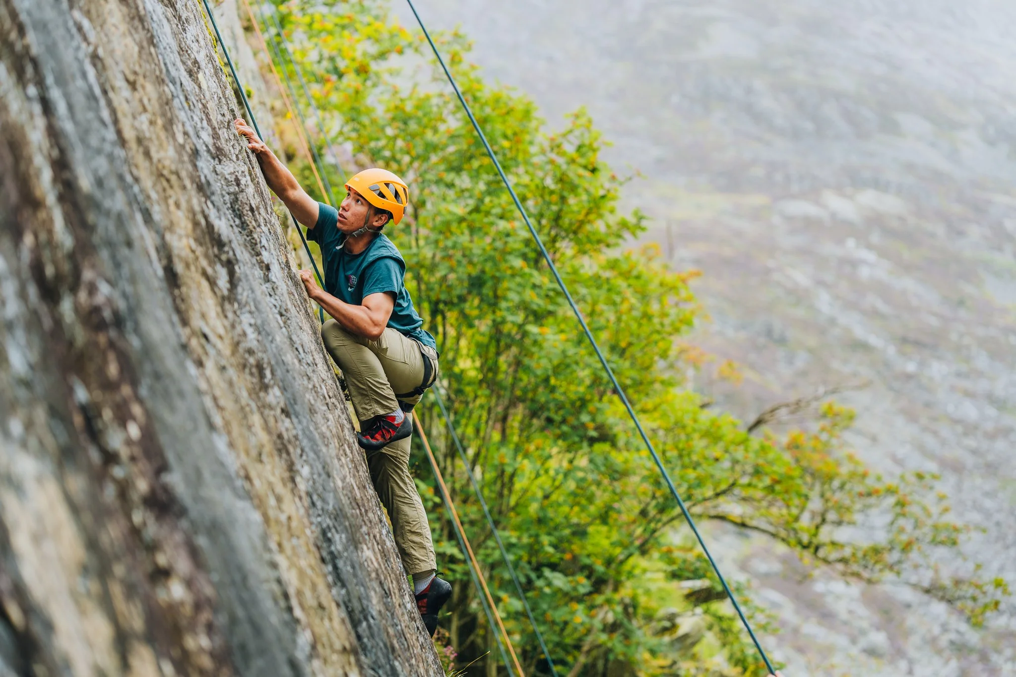 A person wearing a yellow helmet and teal shirt rock climbing a steep cliff with trees and mountains in the background.