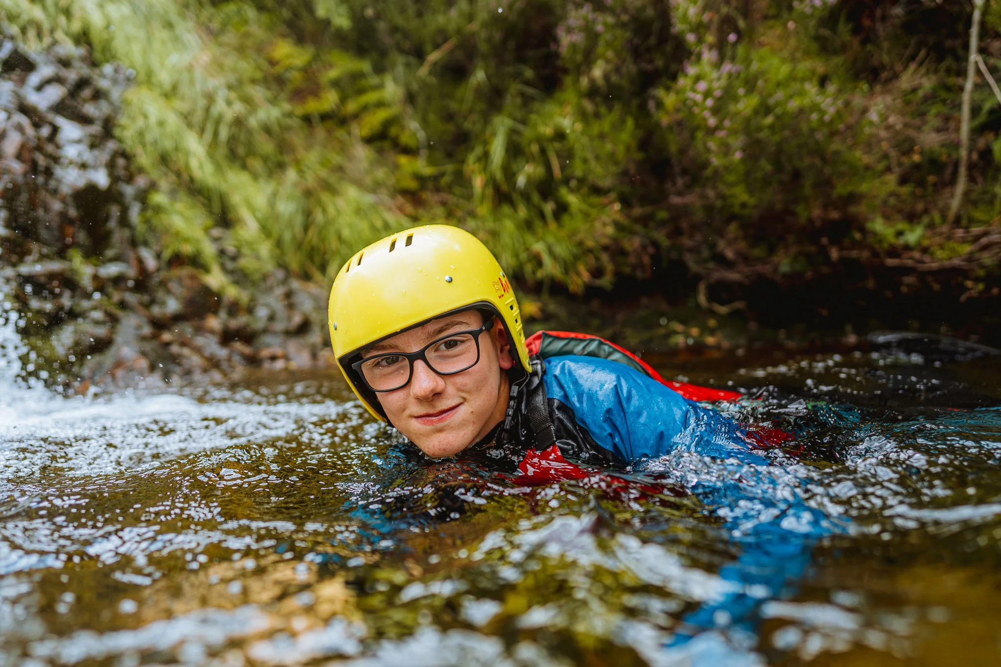 A young person wearing a yellow helmet and blue waterproof gear kayaking in a river surrounded by greenery.