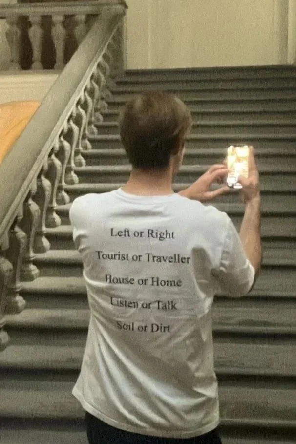 A person with short hair wearing a white T-shirt with instructions to 'Left or Right, Tourist or Traveller, House or Home, Listen or Talk, Soil or Dirt' printed on the back, standing on a staircase, holding a smartphone to take a photo.