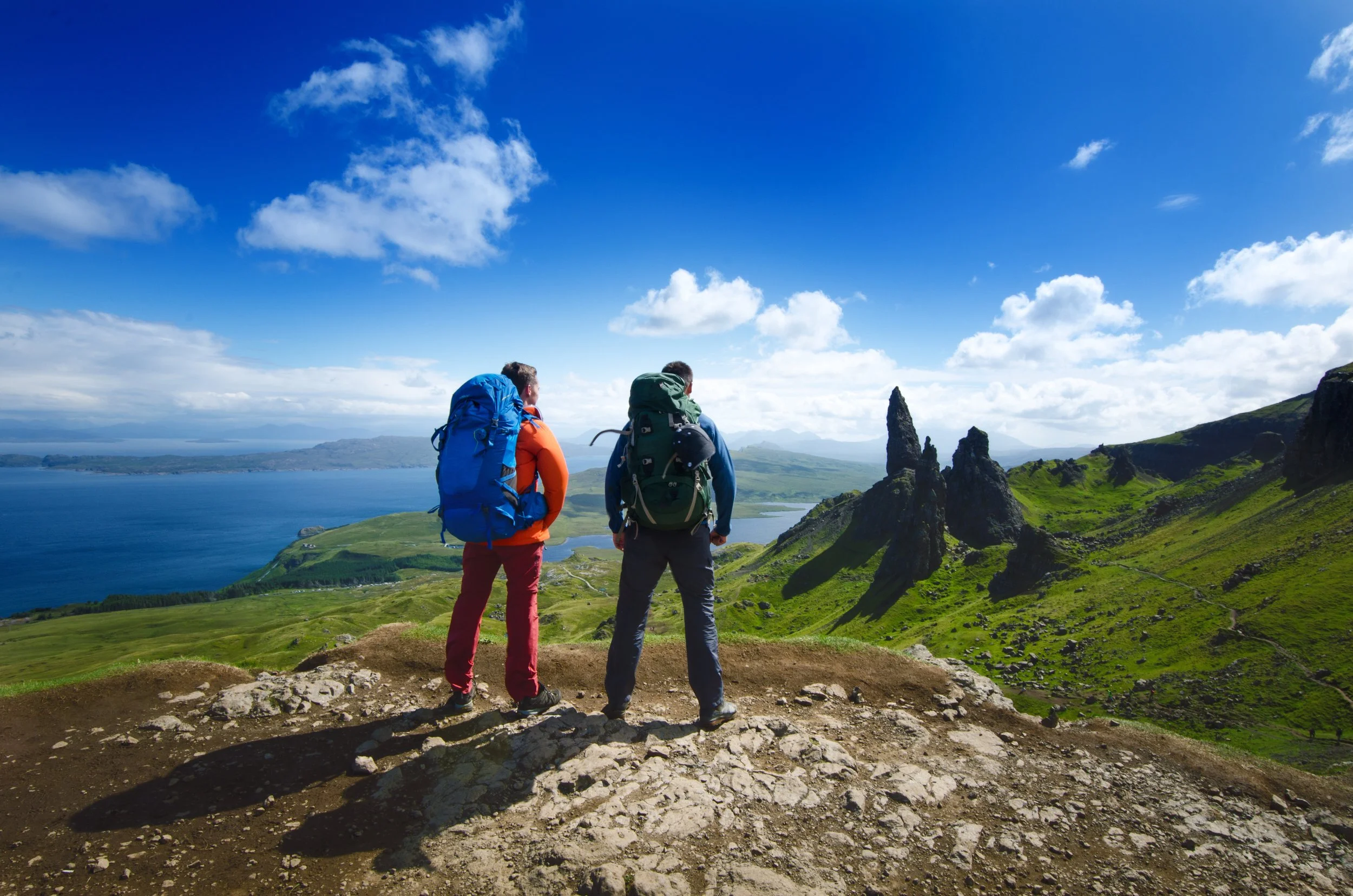Two man admiring the view in front of them, from the top of a hill: green hills, blue lake, blue sky with white clouds