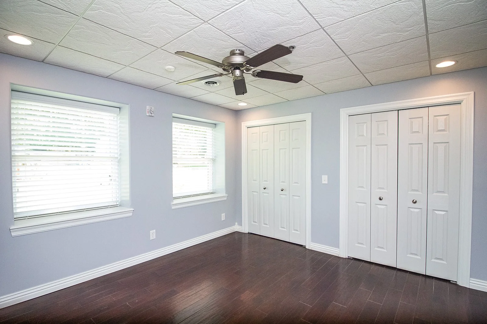 Empty room with light blue walls, two white closet doors, two windows with blinds, a ceiling fan, and dark wooden floors.