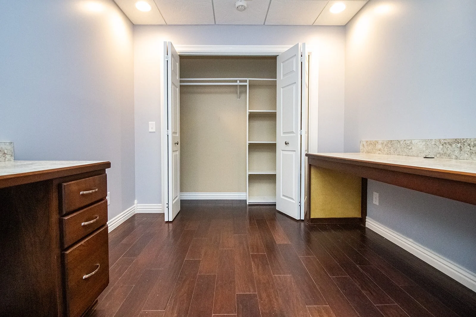 Empty room with hardwood floors, a closet with open double doors, and built-in shelves, with two desks along the sides of the room.