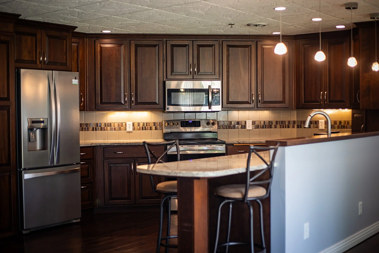 Kitchen with dark wood cabinets, stainless steel refrigerator, microwave, and oven, beige tiled backsplash with decorative border, and a small oval table with two chairs.