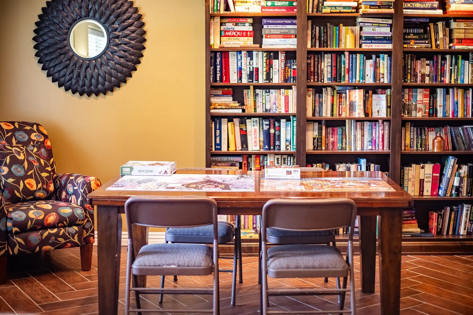 A cozy library room featuring a wooden table with puzzle pieces and a box of 1000 springbok puzzle pieces, two brown chairs, a floral patterned armchair, a large dark wooden bookshelf filled with books, an ornate black sunburst mirror on a yellow wall, and a hardwood floor.