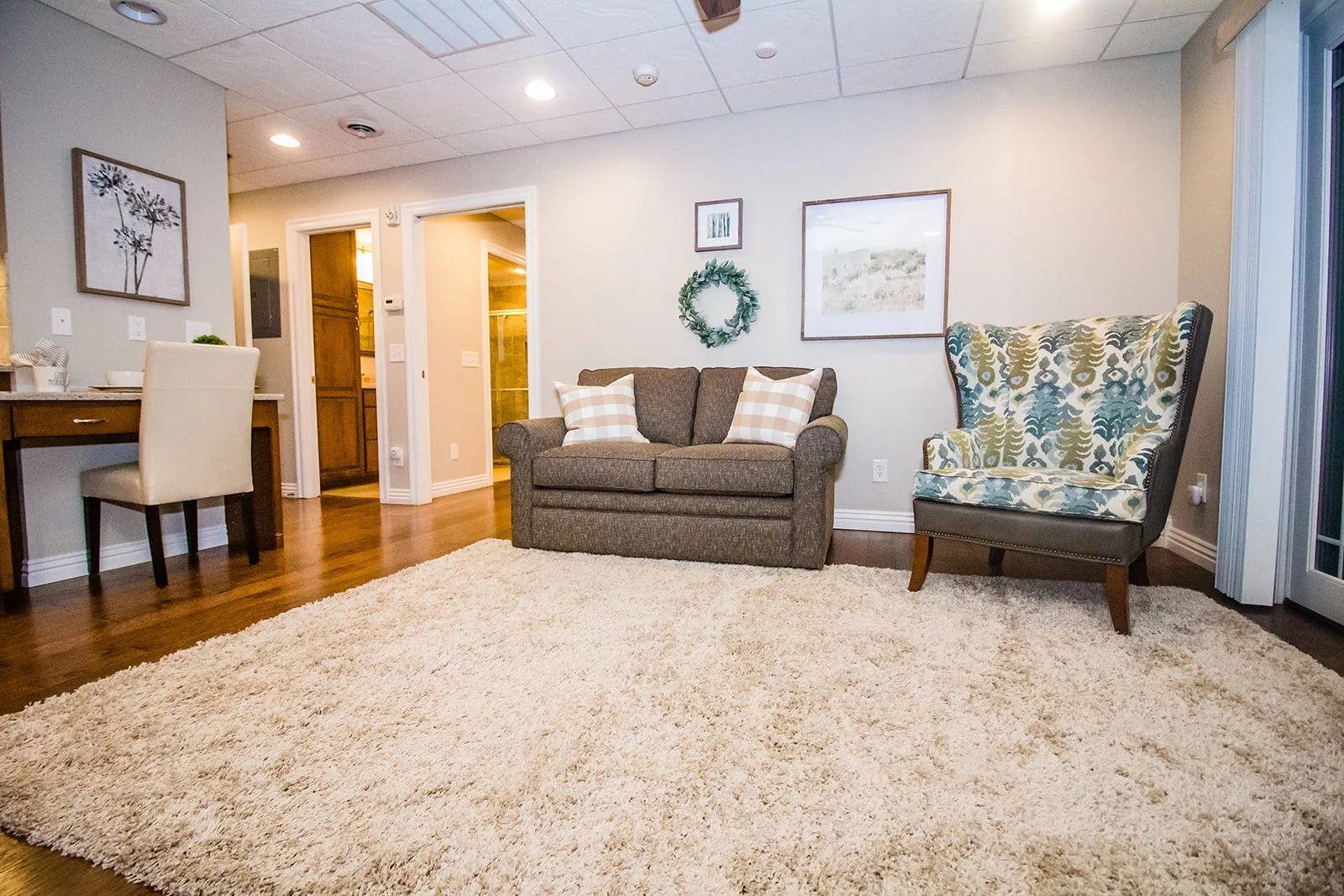 Living room with beige walls, a large cream fluffy rug, a brown upholstered loveseat with checkered pillows, a patterned armchair, and wall art including a wreath and framed pictures.