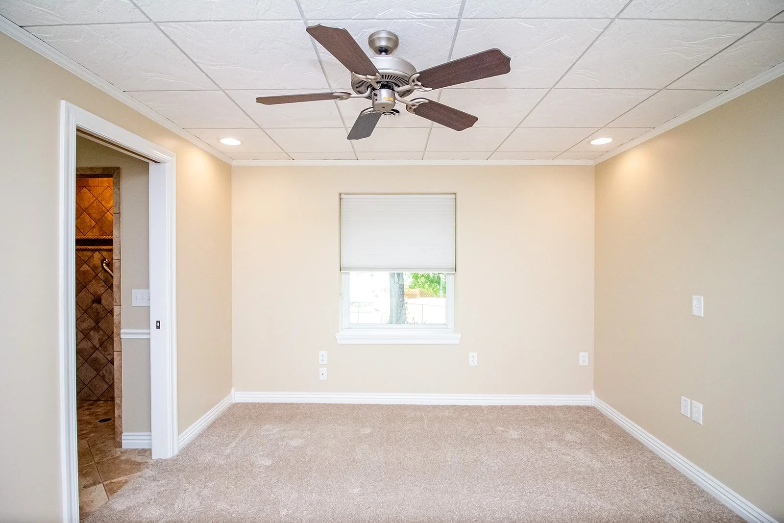 Empty room with beige walls, beige carpet, a window with a closed white blind, ceiling fan with dark blades, and an open door leading to a tiled bathroom.