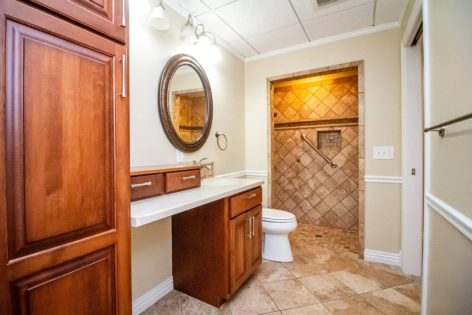 Bathroom with a wooden cabinet, a mirror, a countertop with a sink, a toilet, and a walk-in shower with tiled walls and a grab bar.