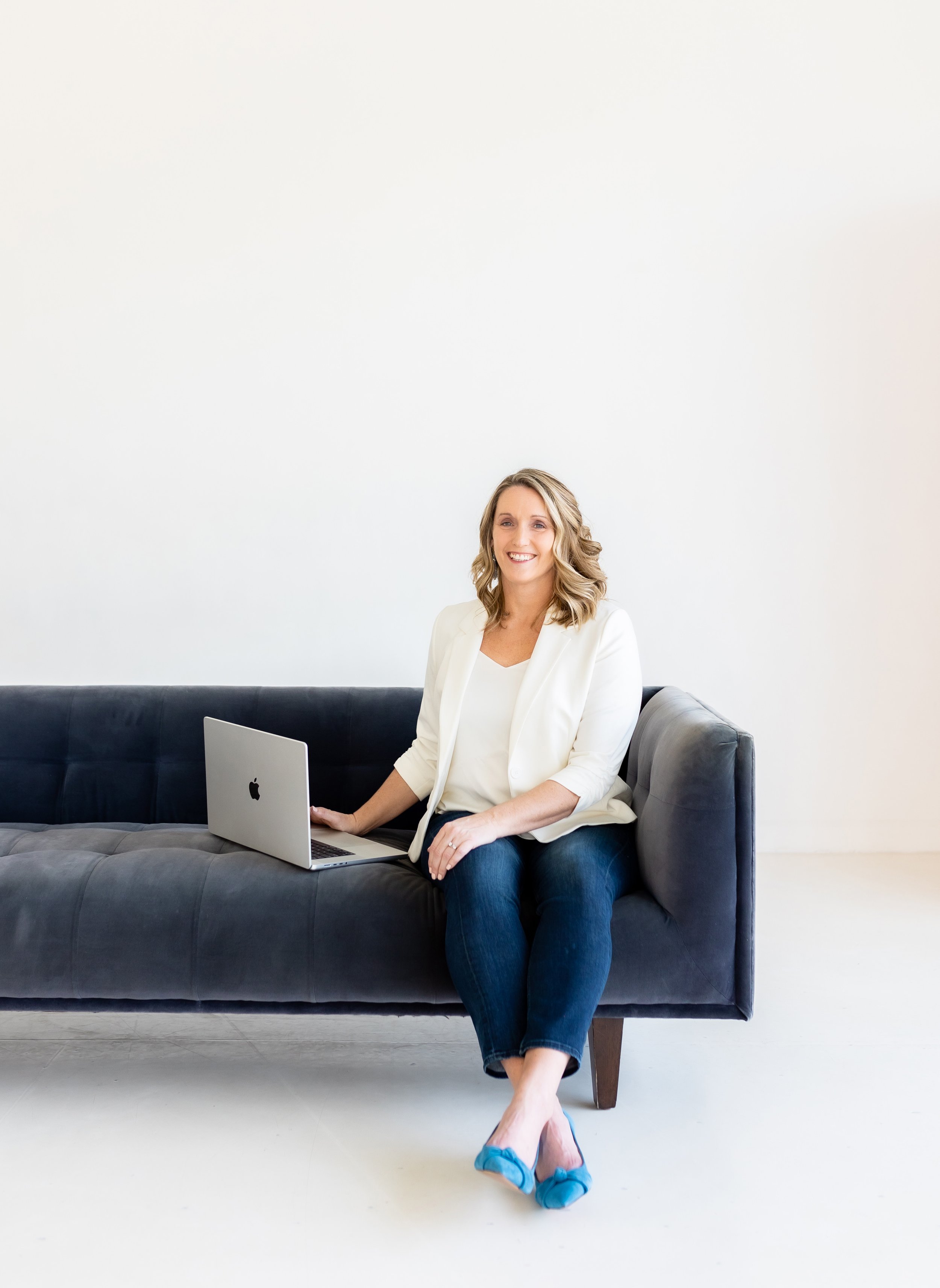 Woman sitting on a dark sofa with a laptop, wearing a white blazer, jeans, and blue flats, against a plain white background.