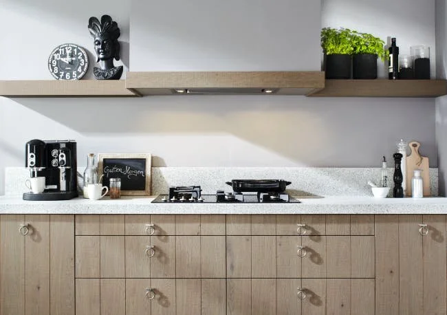 Modern kitchen with wooden cabinets, gas stove, espresso machine, wall clock, small chalkboard, and shelf with plants and kitchen items.