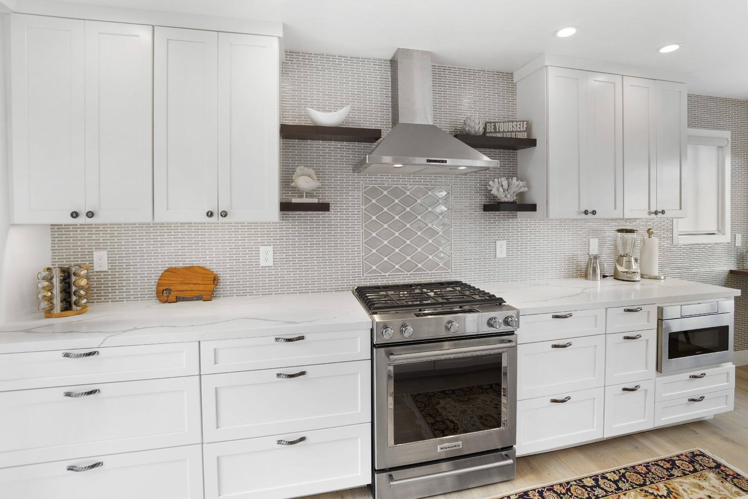 Modern kitchen with white cabinets, stainless steel stove, and range hood, featuring a mosaic tile backsplash, open shelves, and countertop appliances.