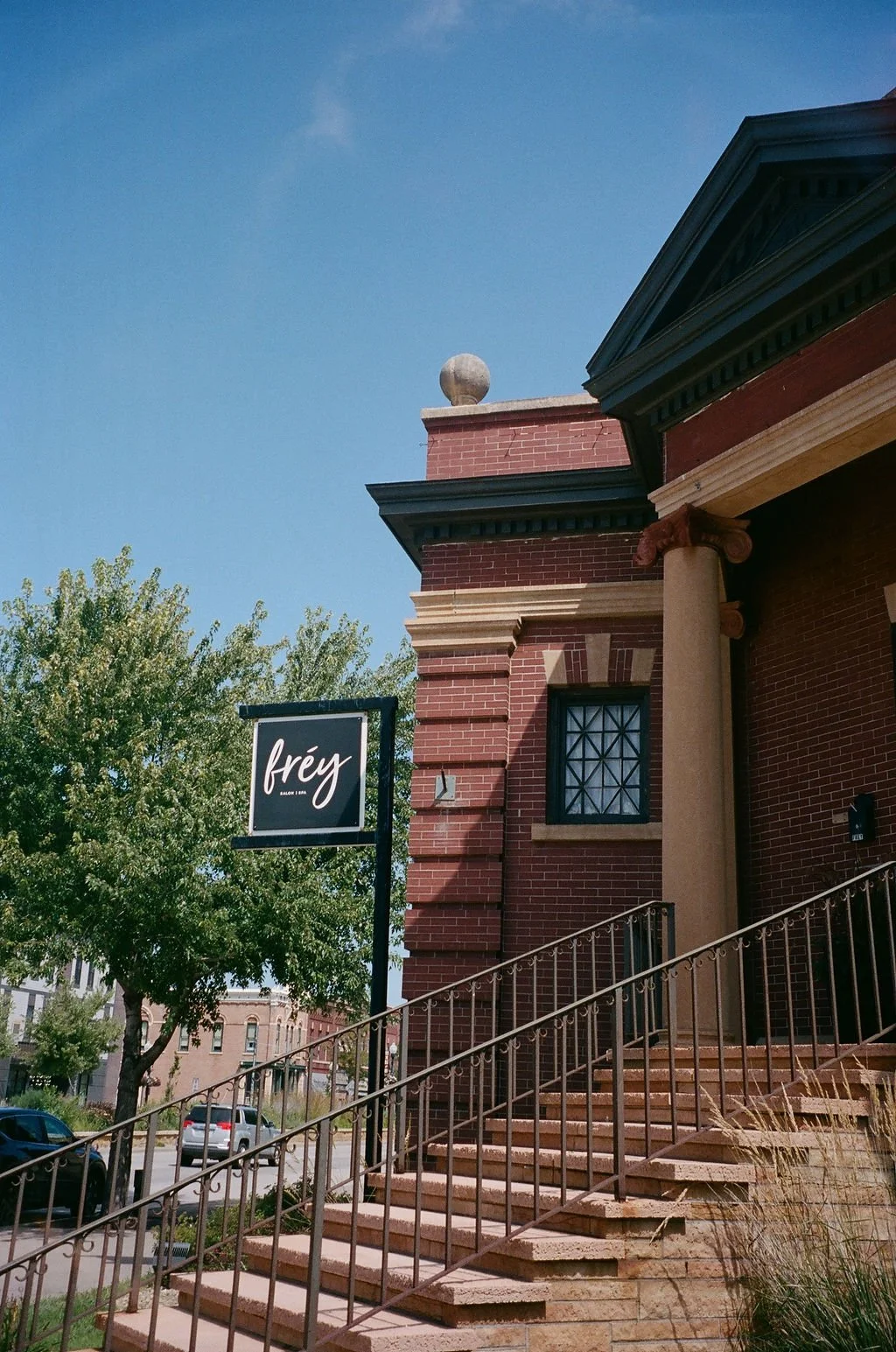 A brick building with stairs leading up to a door, a sign reading 'frey,' and a tree in a sunny urban setting.