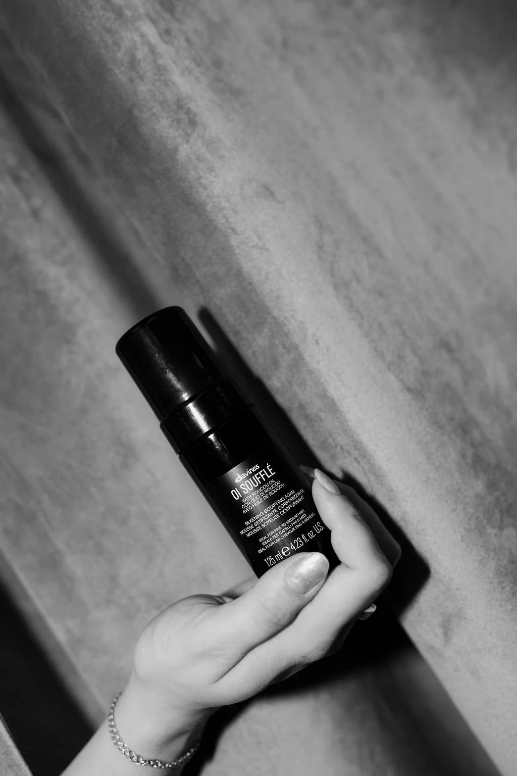 Hand holding a small black bottle labeled 'Davines OI SOUFFLE' against a textured surface.