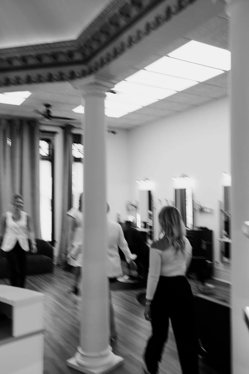 Black and white photo of a room with a high ceiling and ornate crown molding, people standing and talking, a large window with curtains, and furniture like a piano and lamps.