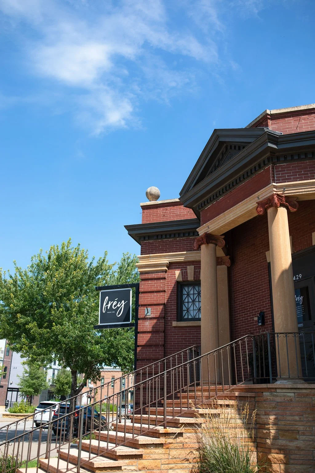 Exterior of a brick building with classical columns, stairs, and a sign that reads 'fréy' under a clear blue sky with a few clouds.