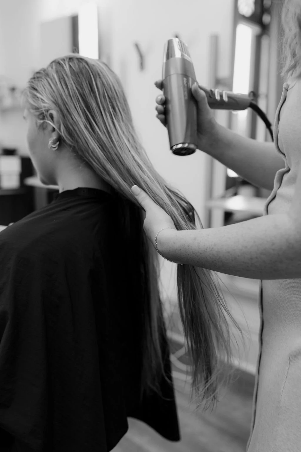 A woman is getting her hair blow-dried by a stylist in a salon. The stylist is using a blow dryer to style the woman's long, straight hair.