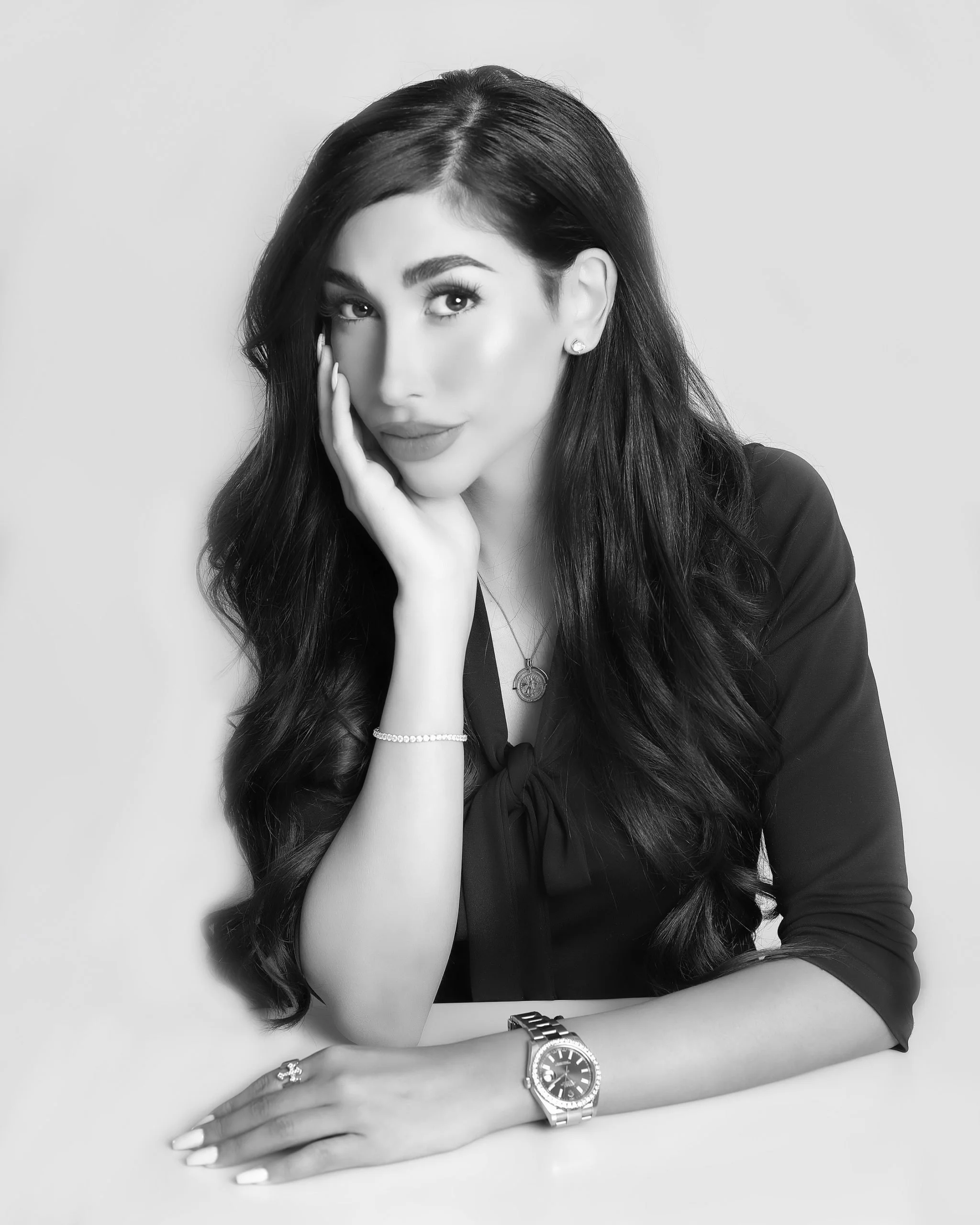 Black and white portrait of a woman with long wavy hair, wearing a dark blouse, a watch, necklace, earrings, and a bracelet, resting her chin on her hand with a slight smile.