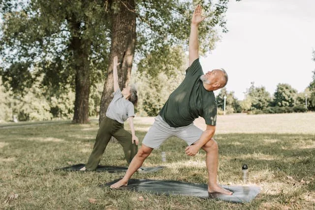 Two people practicing yoga outdoors in a park, one adult man and one elderly woman, both in yoga poses on yoga mats with trees and grass in the background.